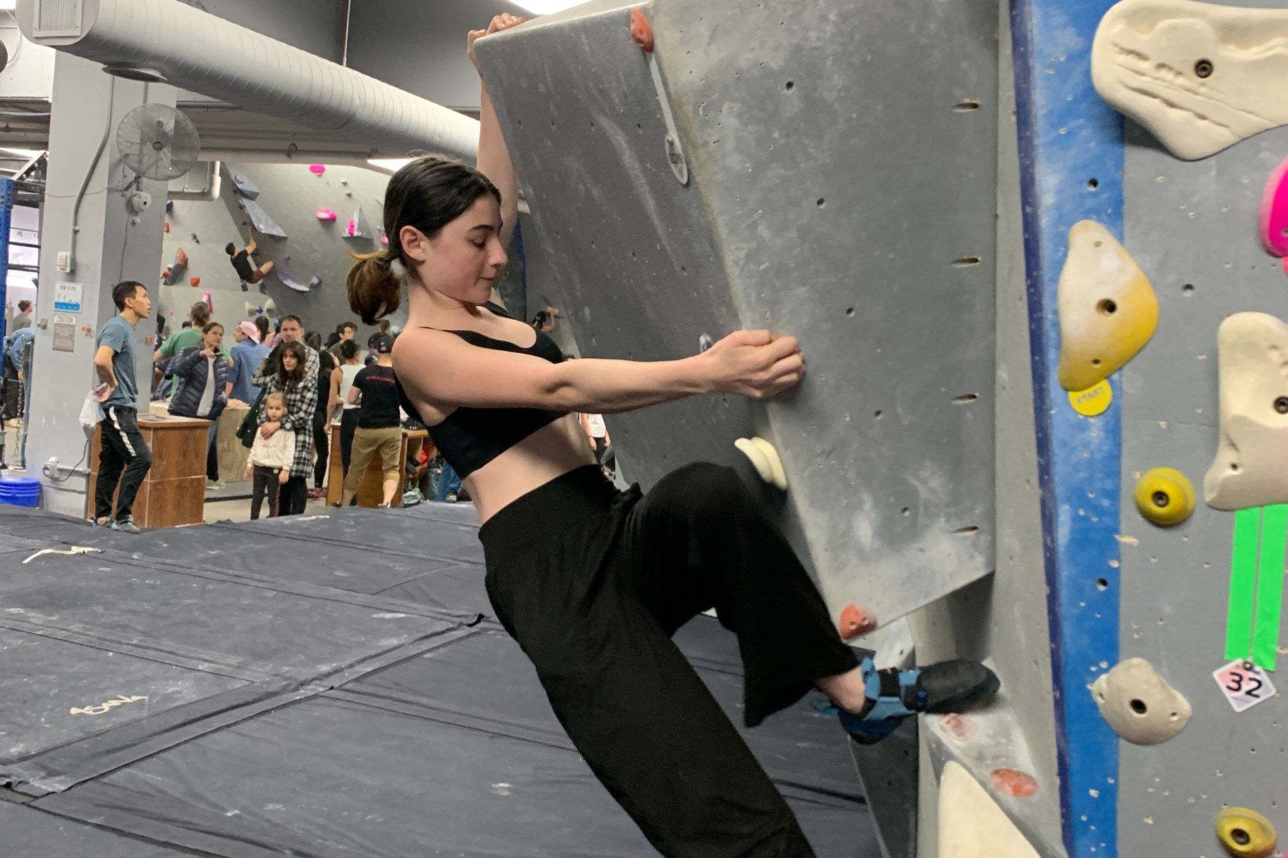 A teen girl holds onto blocky volumes and steps her foot up on the bouldering wall.