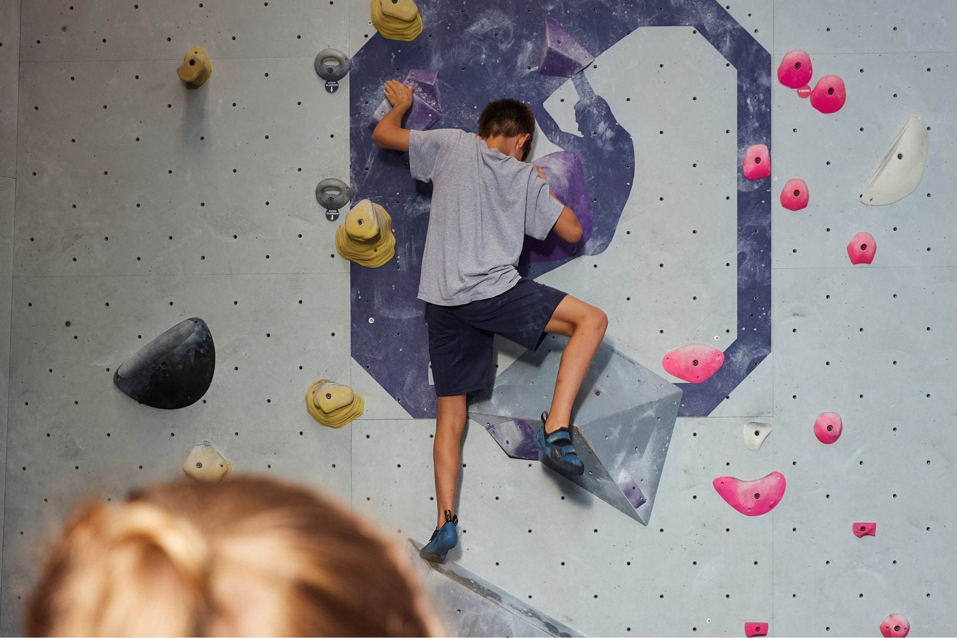 A boy is bouldering on a climb with block shaped slopey holds. The back of a girl's head is visible and the CRG logo (the silhouette of a climber) is on the climbing wall.