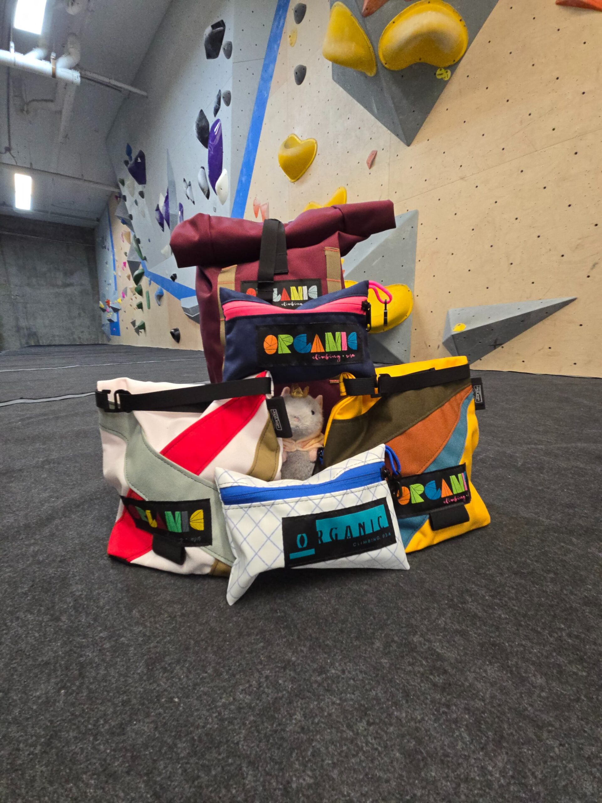 A stack of organic chalk bags and products arranged in front of a bouldering wall at Central Rock Chelsea. In the middle of the chalk bags is a stuffed rat wearing a crown and cape.