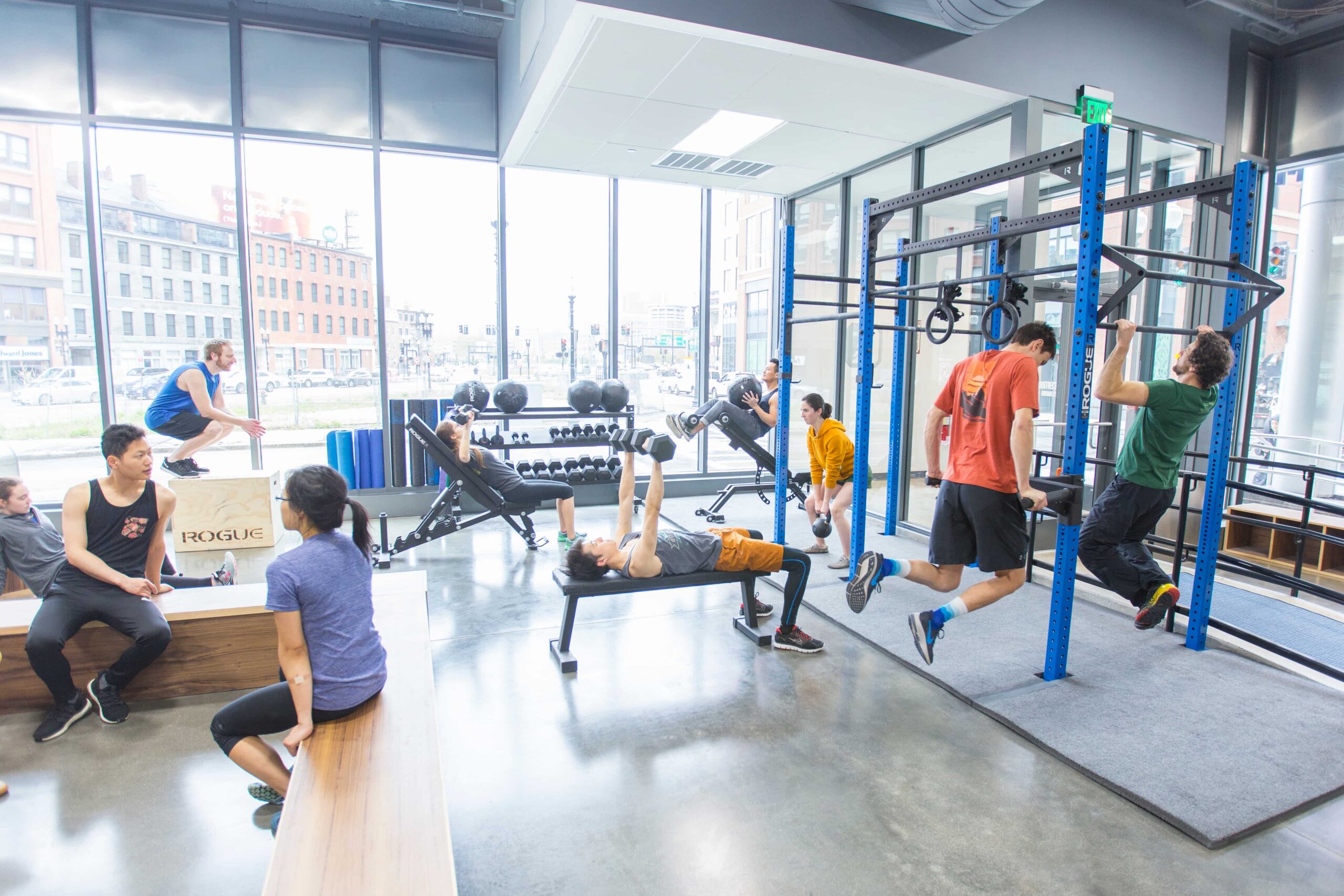 Members work out in the fitness area at Central Rock Gym North Station — doing pull-ups, squatting, bench pressing, dips, and box jumps with floor-to-ceiling windows overlooking Boston