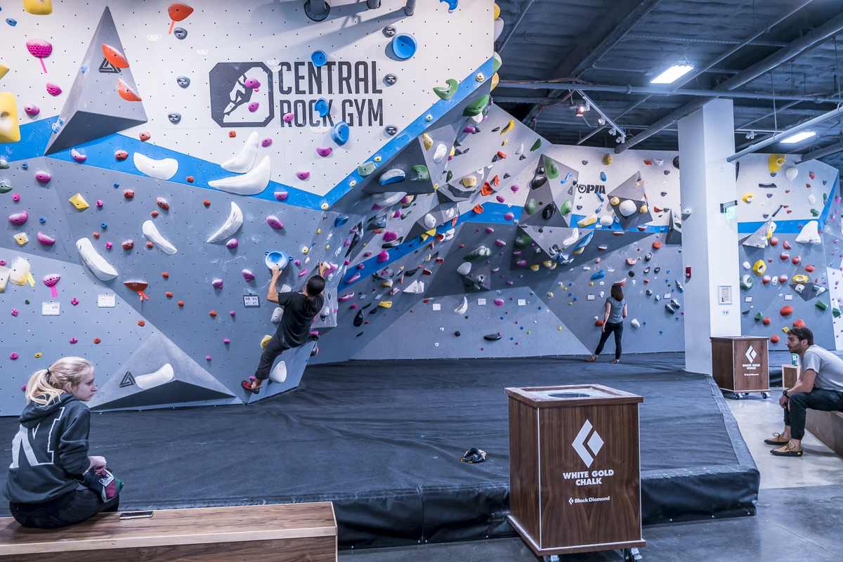 A man locks off on the bouldering wall at Central Rock Gym North Station while several people sit on a bench and watch, with the Central Rock Gym logo visible on the wall