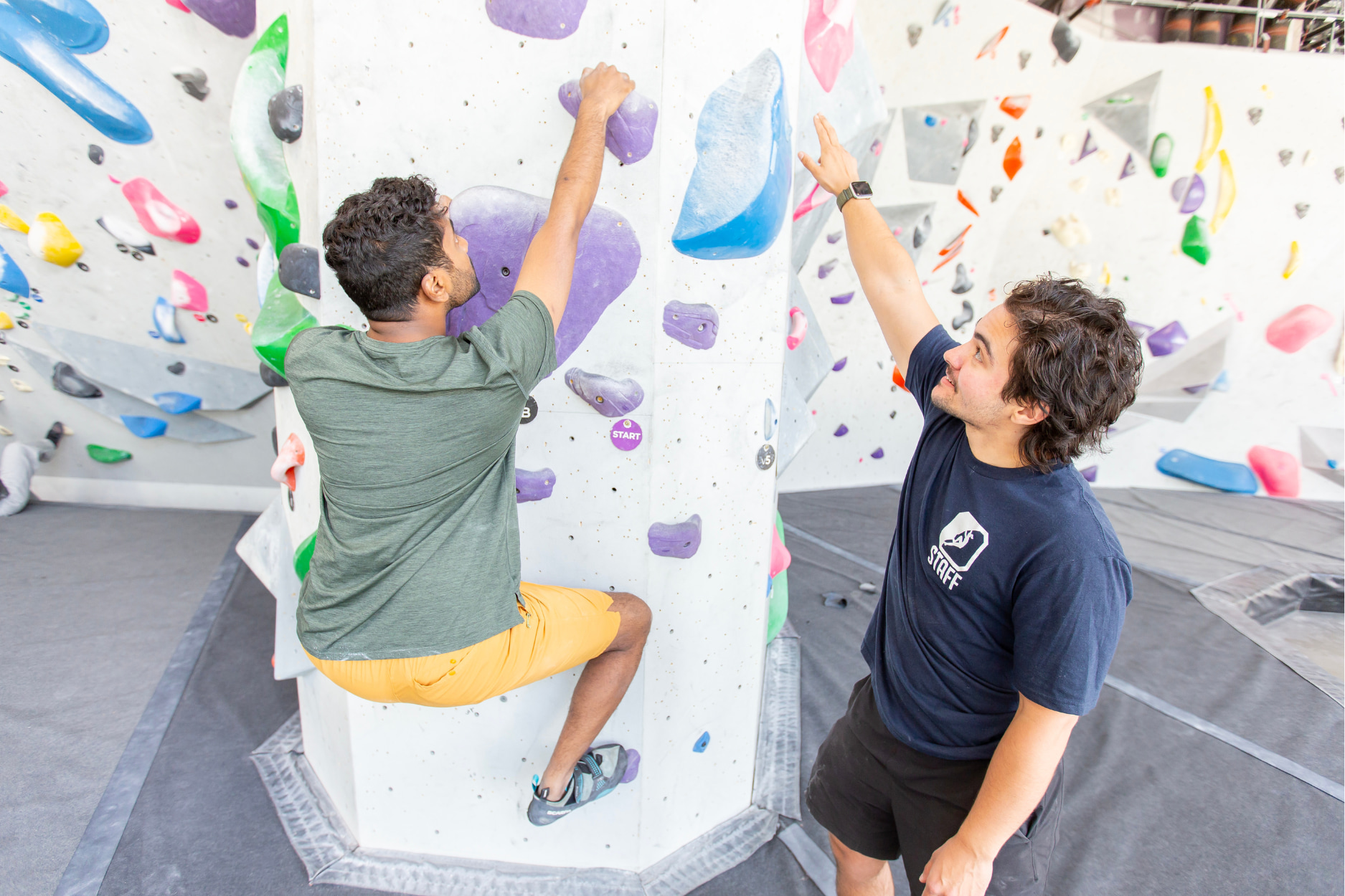 A man who works at Central Rock Gym is teaching a beginner about rock climbing and showing pointing out the next hold to him on the bouldering wall.