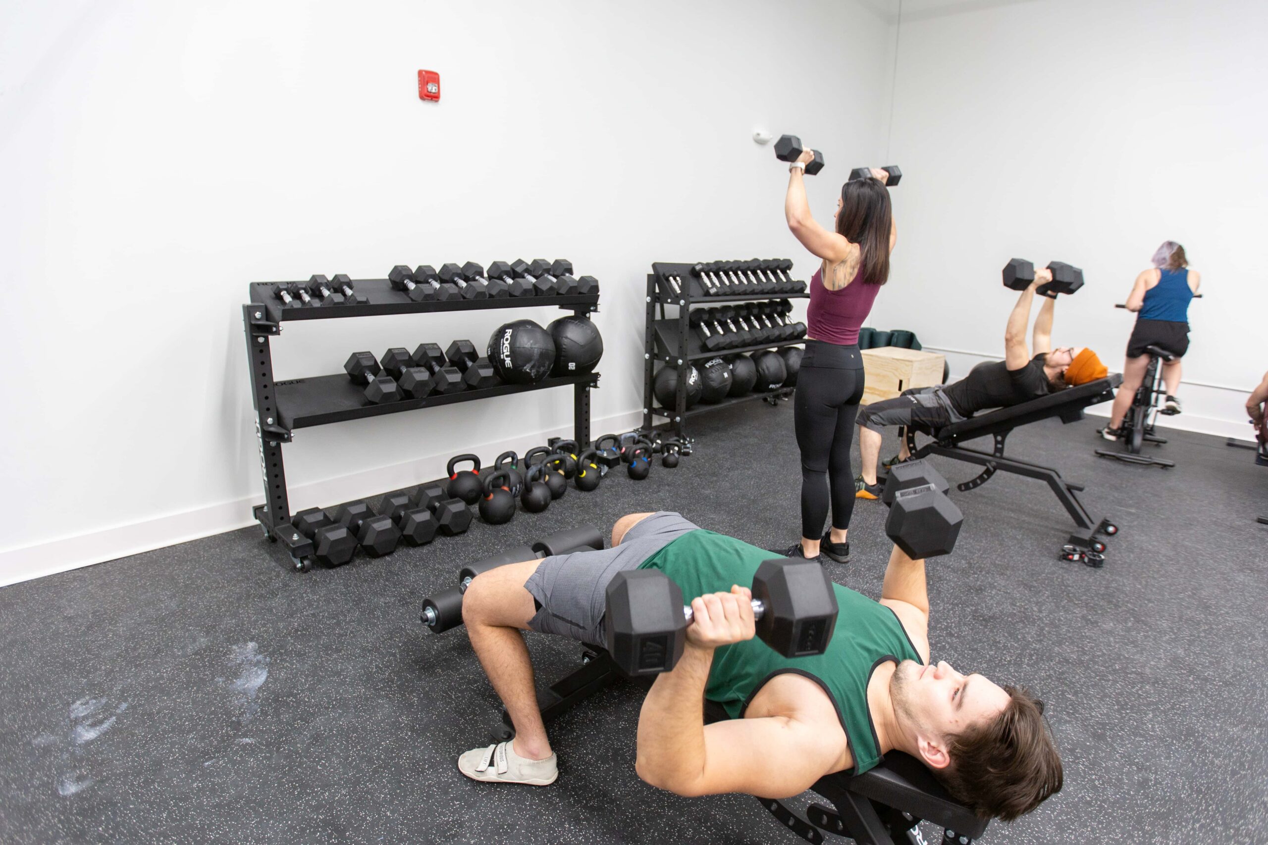 A man presses dumbbells while other climbers workout in the background.