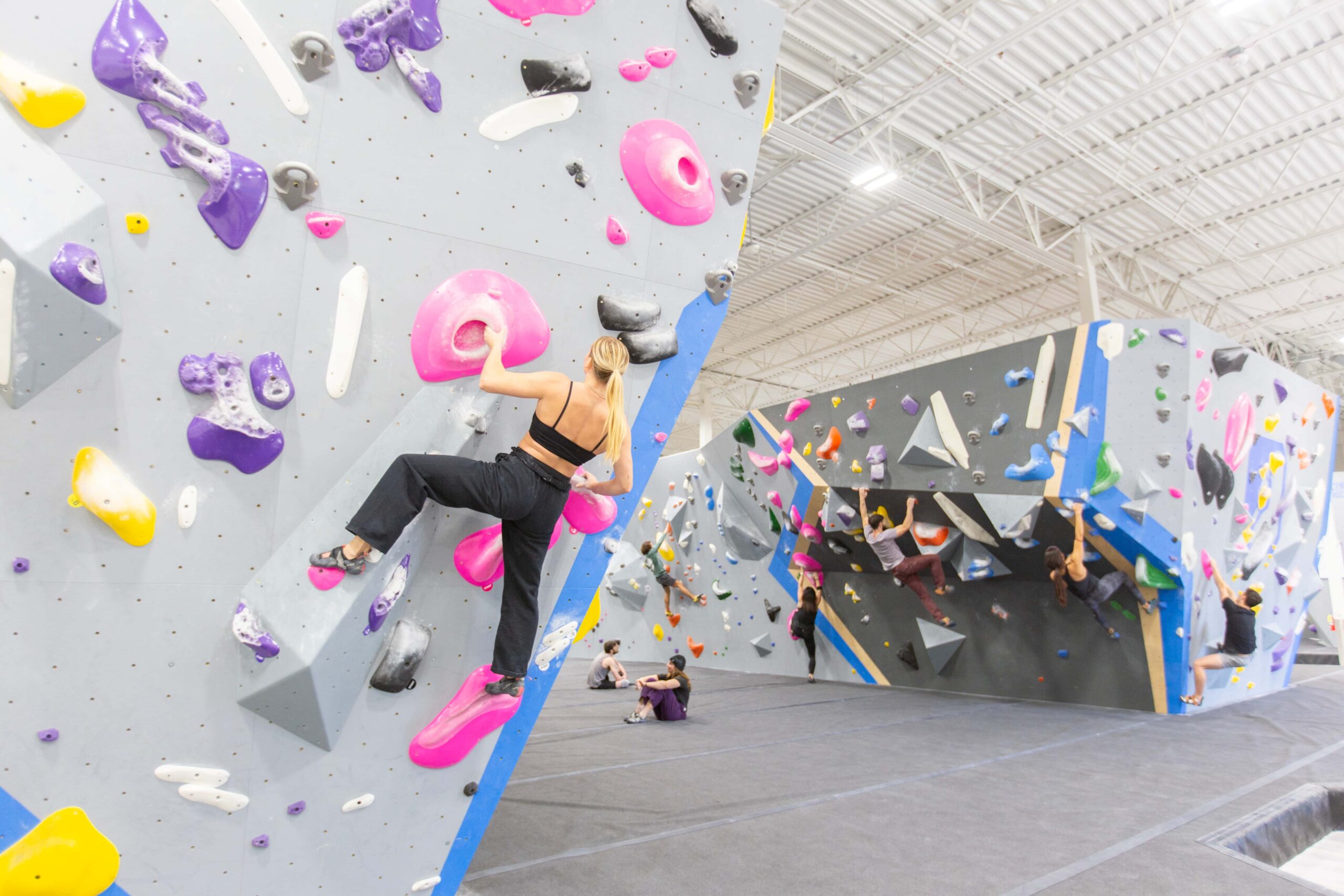 A girl holds onto a jug on the climb. In the background boulderers climb a steep wall.
