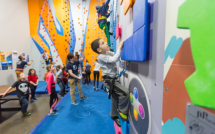 A young child climbs a colorful indoor rock wall, secured with a harness. Other children watch, some with excitement, in a lively, bright play area.