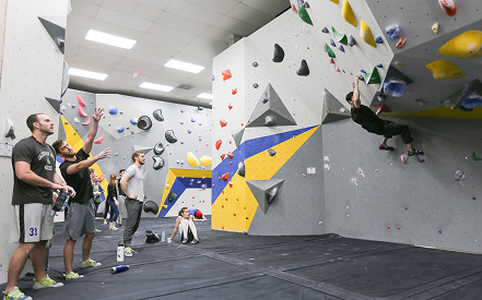 A group of people at an indoor climbing gym watch and cheer as a climber ascends a textured wall with colorful holds. The atmosphere is supportive.