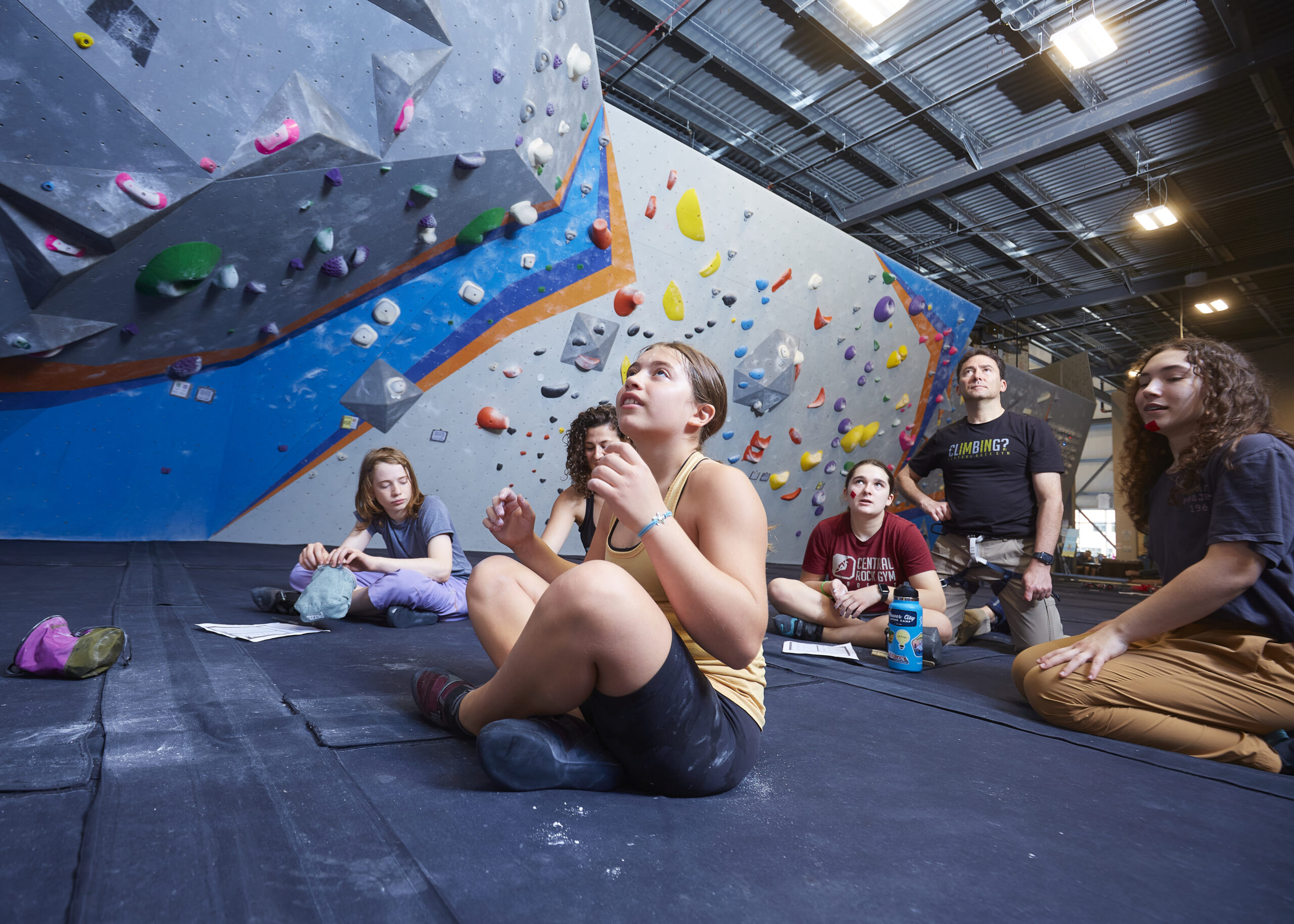 Kids sit criss cross on the mats with their climbing coaches as they sequence a boulder.