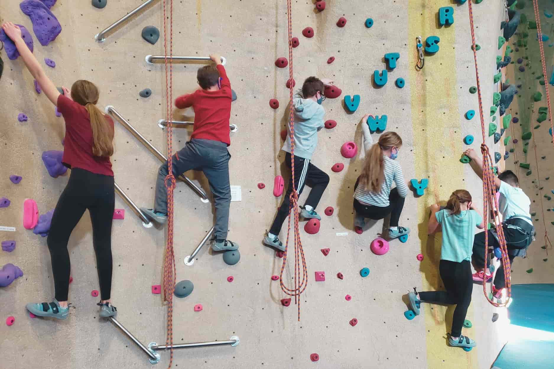 A group of 6 children climbs on a climbing wall at Central Rock gym. Two of the children with the red shirts are climbing upwards , and the other 4 are climb towards the left side of the wall.