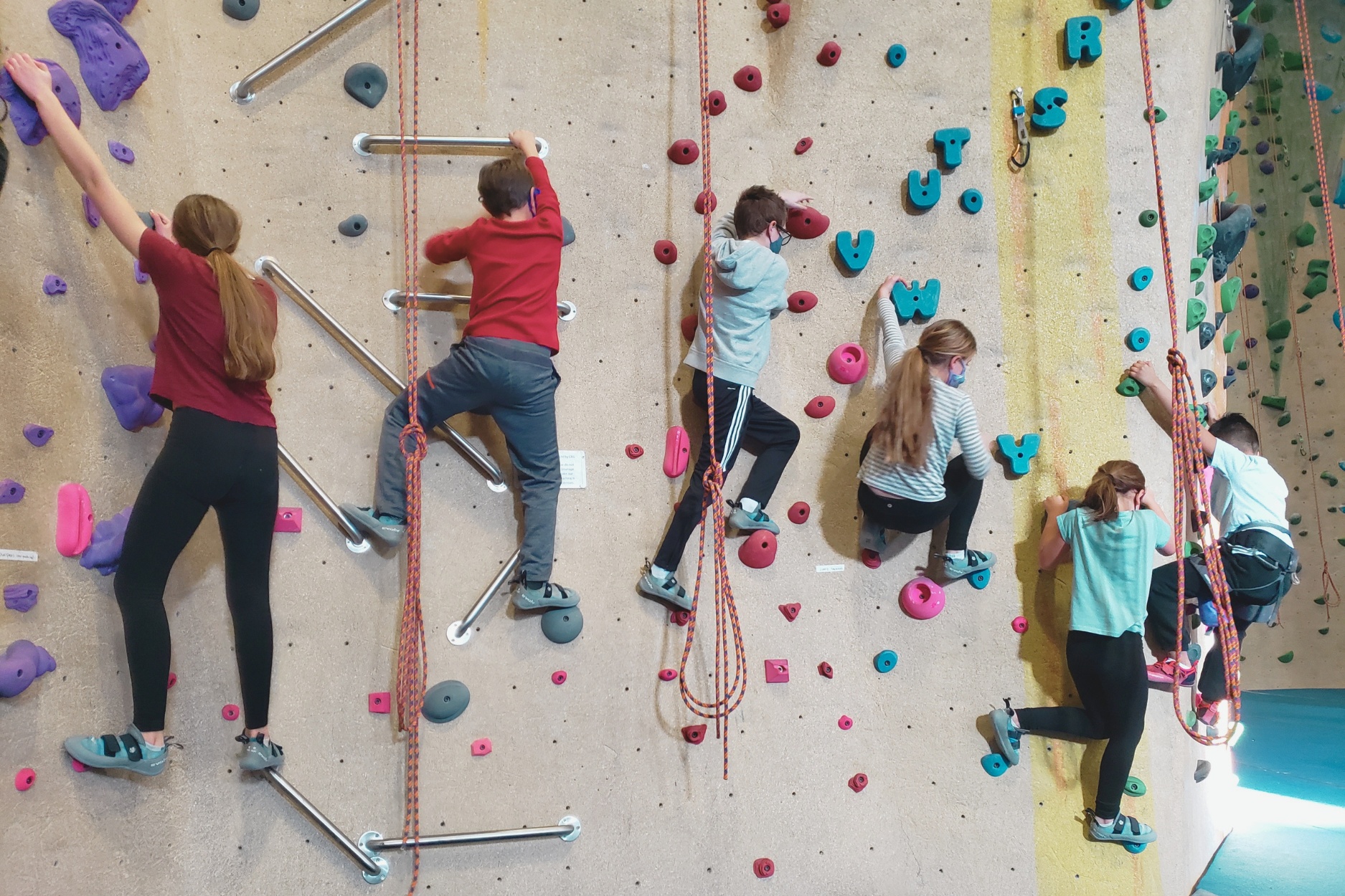 A group of 6 children climbs on a climbing wall at Central Rock gym. Two of the children with the red shirts are climbing upwards , and the other 4 are climb towards the left side of the wall.