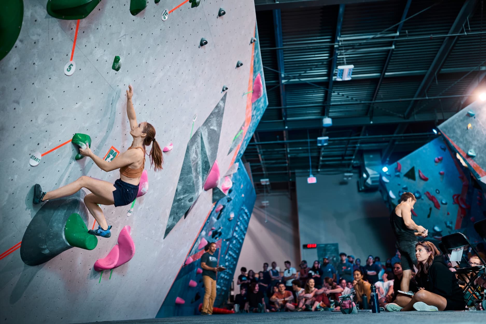 Female bouldering on an overhanging wall angle in the Canal Crusher finals with crowd seen in the background