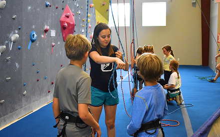 A female instructor teaches two boys wearing harnesses about climbing in an indoor gym. A colorful climbing wall and seated children are visible.