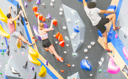Three people bouldering on a colorful indoor climbing wall. They grip vibrant handholds with focus and determination against a geometric backdrop.