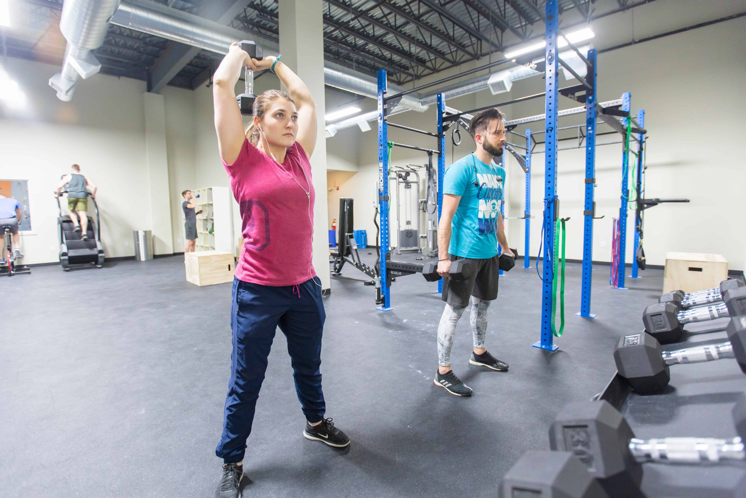 A woman does overhead triceps extensions while a man beside her does dumbbell curls.