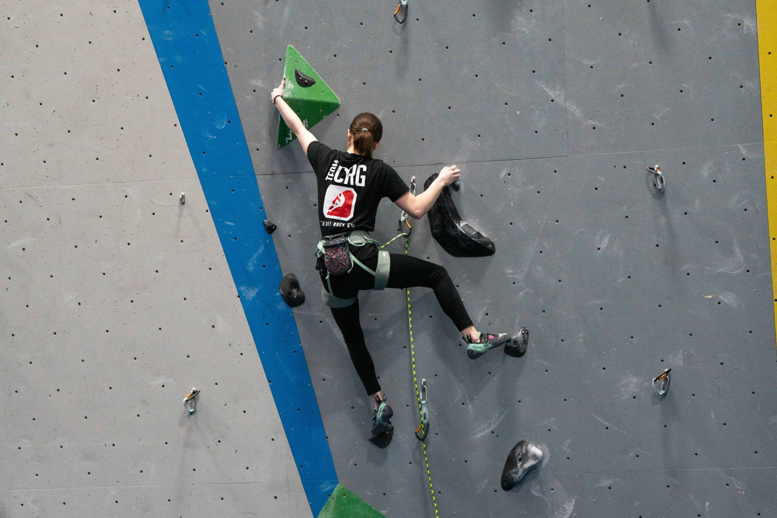 Teenager girl in black CRG Stoneham team t-shirt lead climbs a black route with green volumes