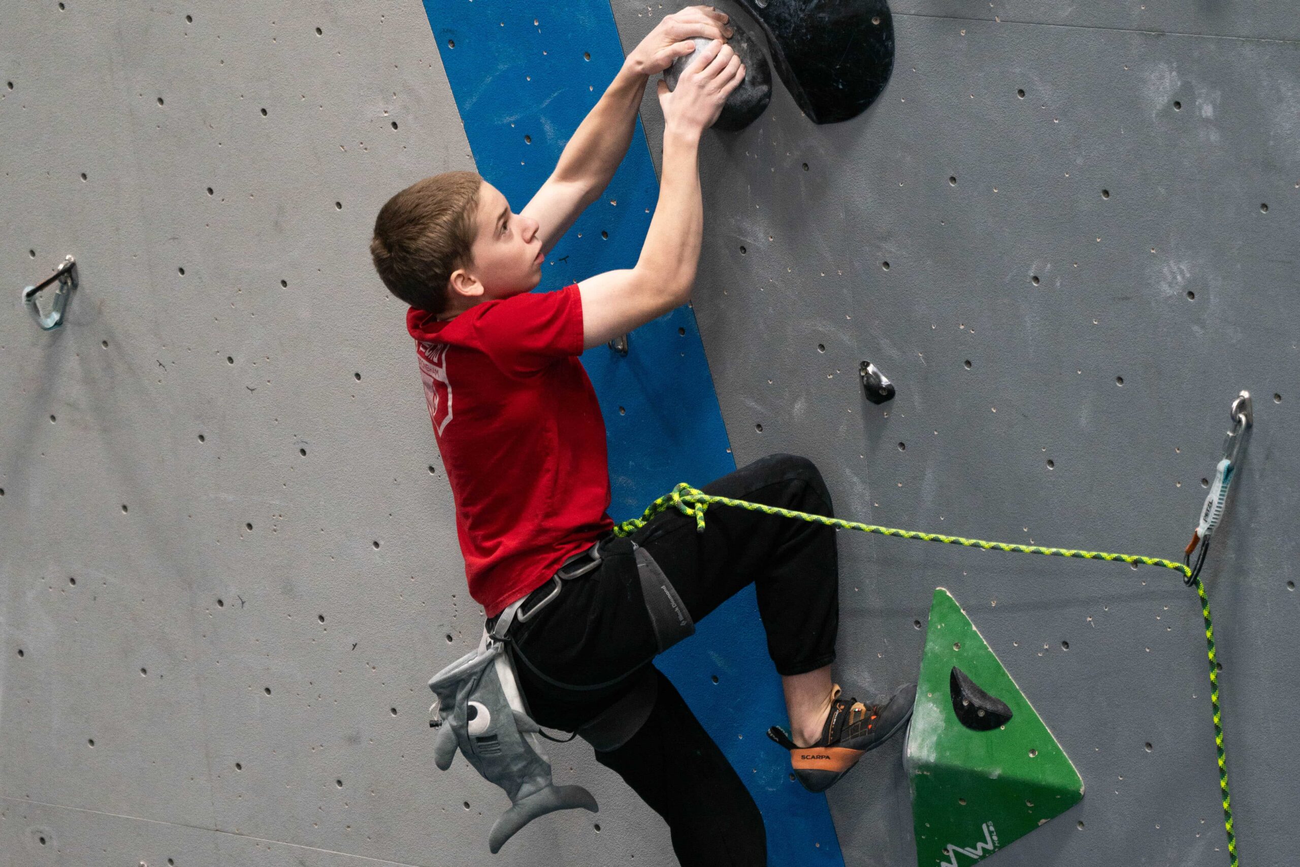 Pre-teen boy in a red CRG Stoneham jersey lead climbs a black route with green volumes