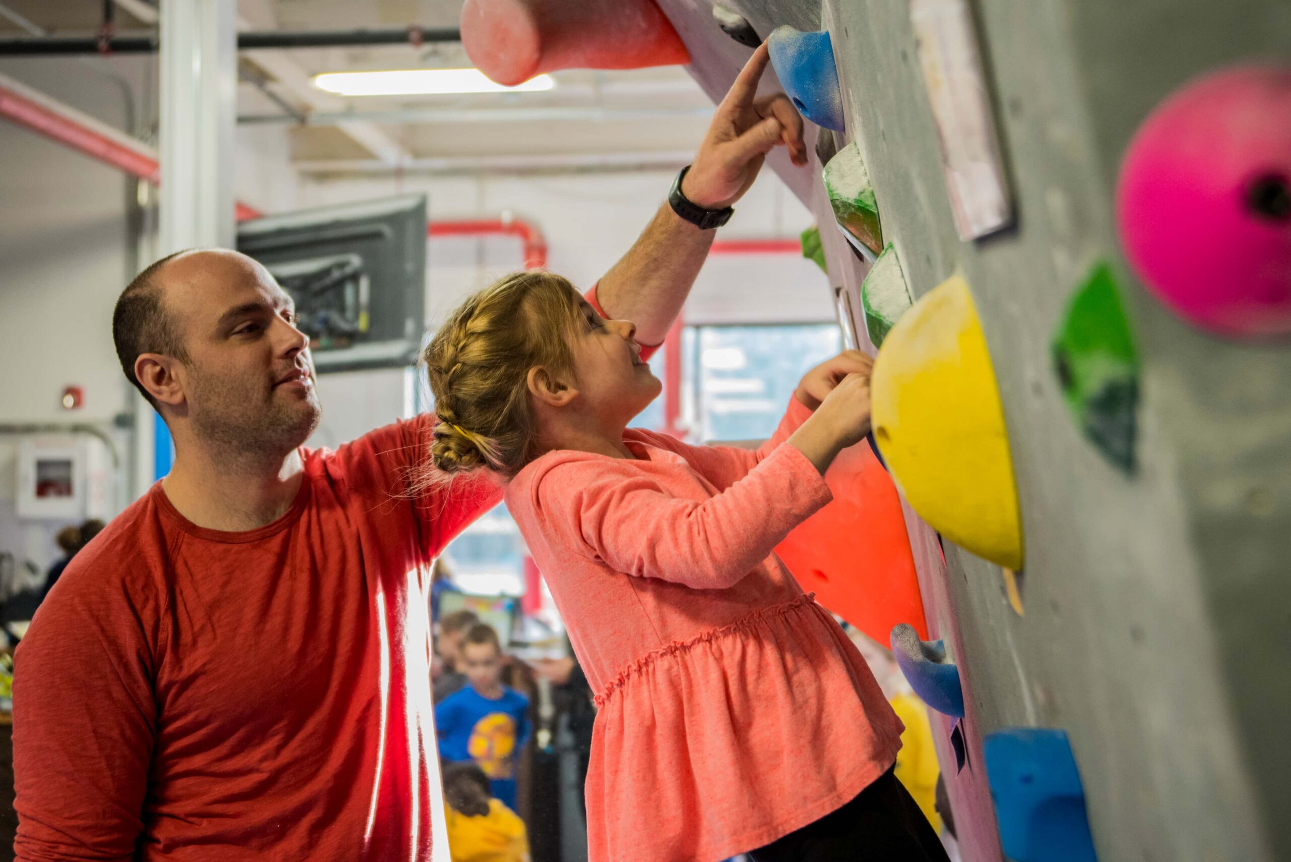 A father points out the next hold to his toddler as she boulders.