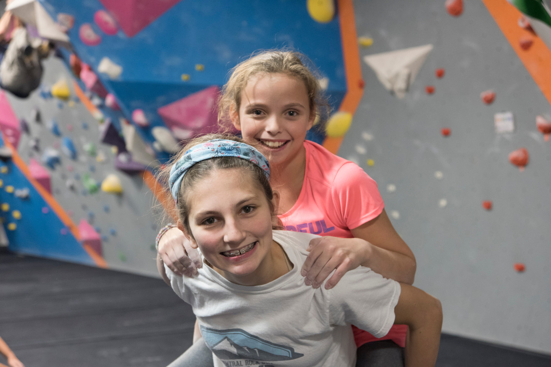 A young girl with braces and a headband gives her friend a piggy back ride.