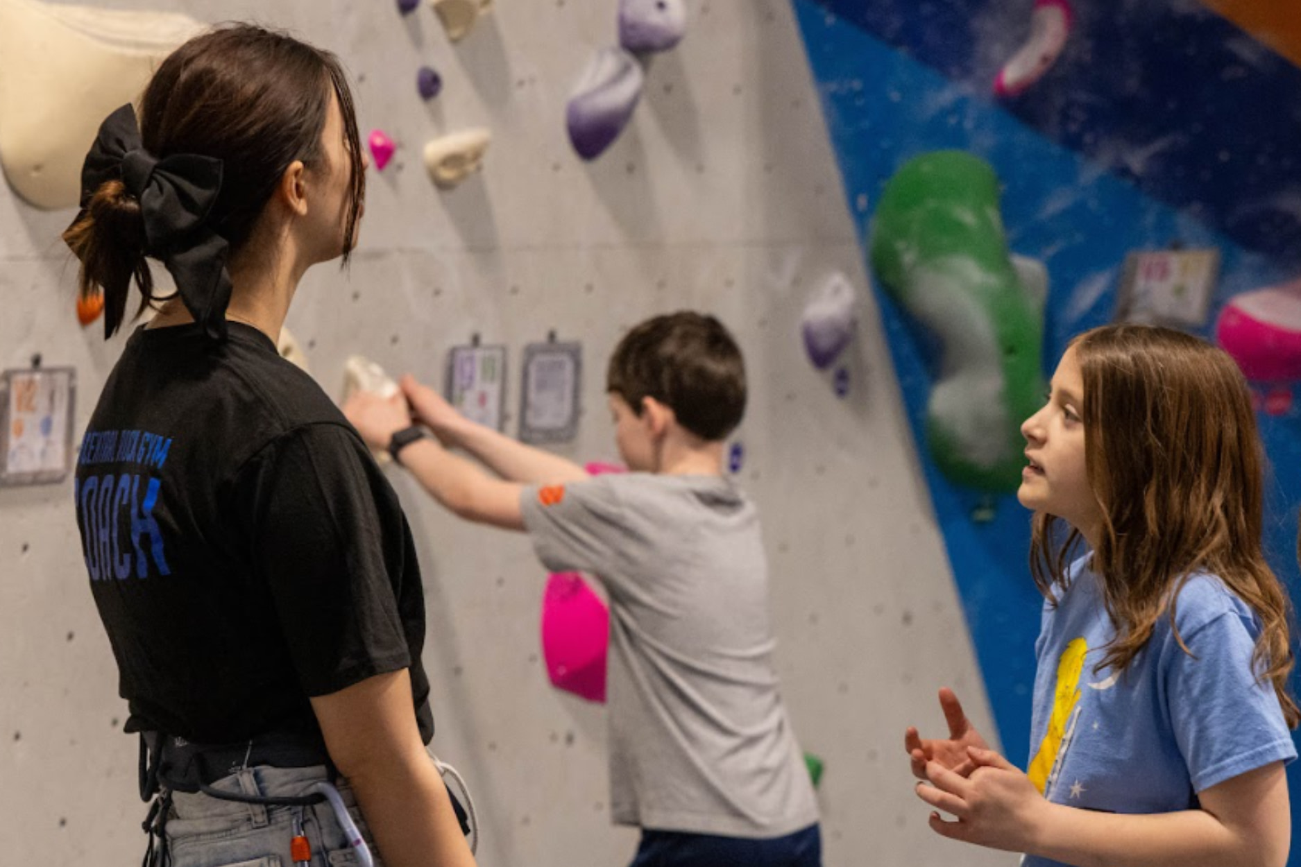 A young girl talks with a climbing coach while a boy boulders in the background.