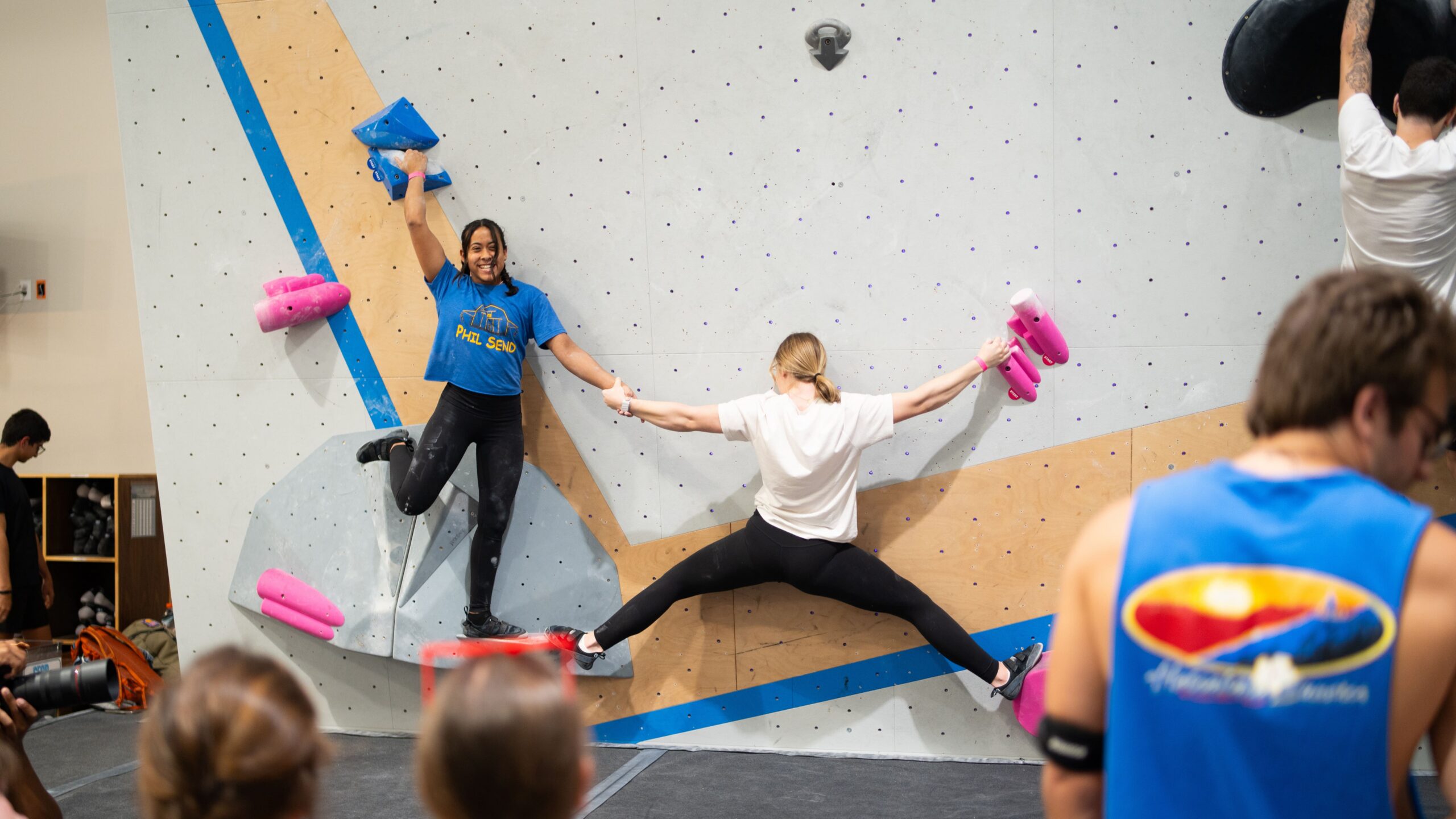 Two girls hold hands as they climb a partner climb together. One girl has her arms and legs stretched out while the other girl is hanging on tight with her back to the wall and is holding onto a jug above her head.