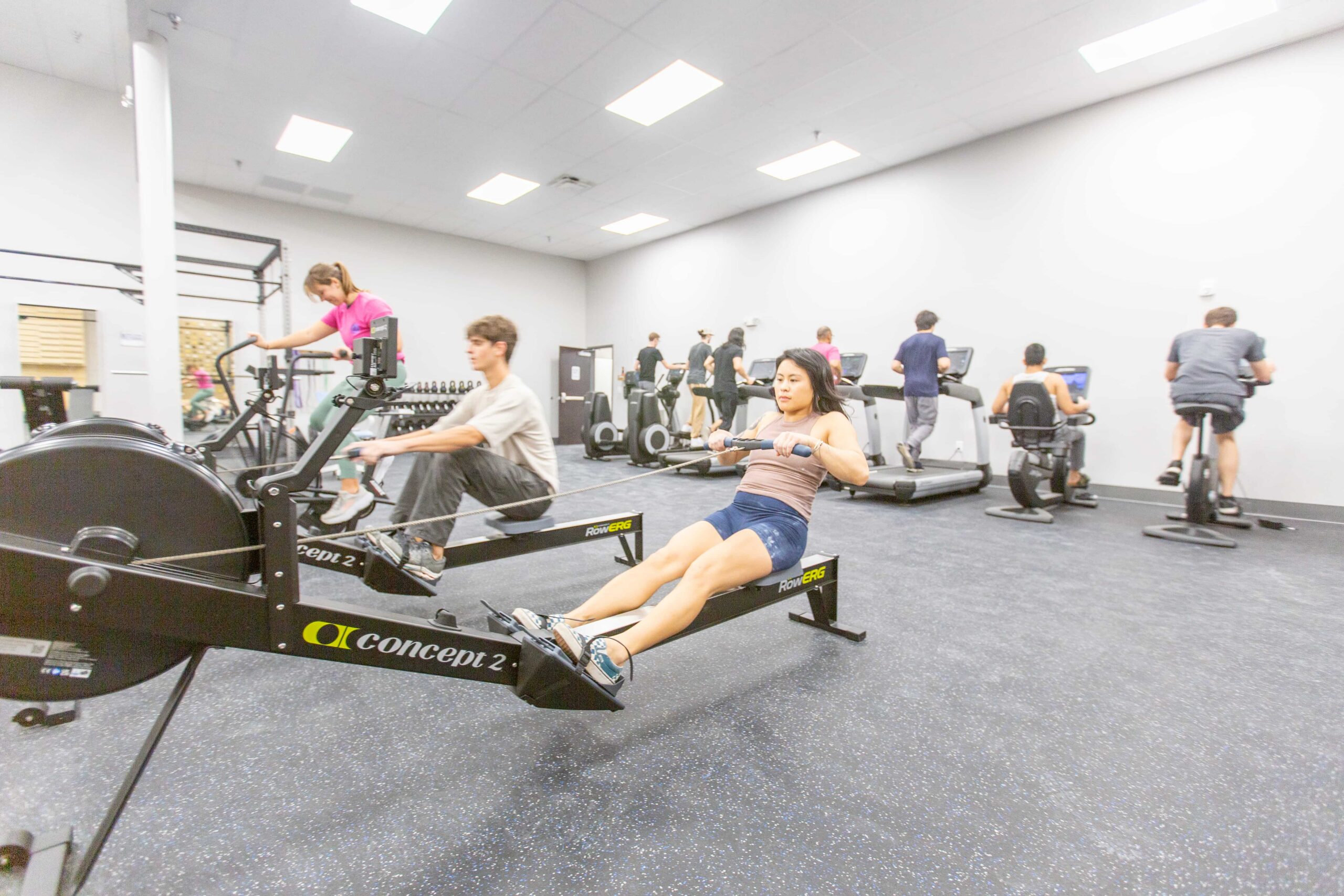 a woman is on a rowing machine. Behind her others workout on various cardio machines.