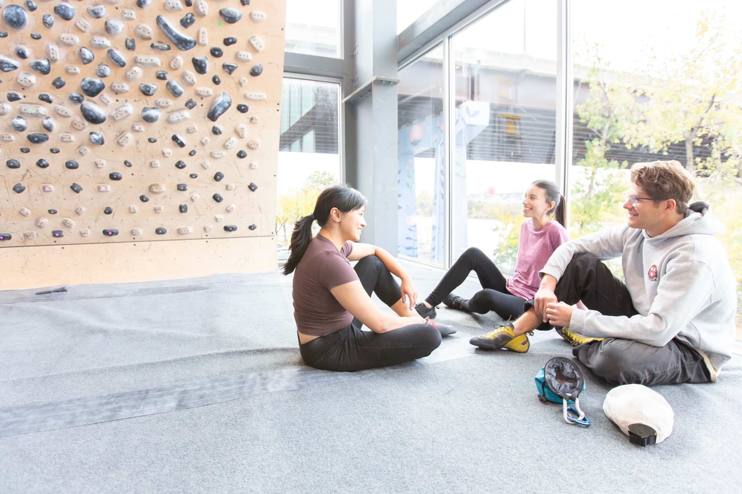 Climbers sit and chat on the mat underneath the Tension board. Outside the window is the Collar City Bridge