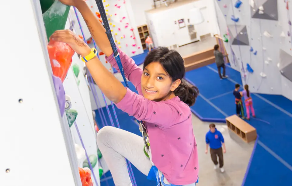 A woman in a pink star-print hoodie top-rope climbs, her leg stepped high on the wall, harness and belay device visible at her waist. A man in a blue hoodie walks across the gym floor below at Central Rock Gym