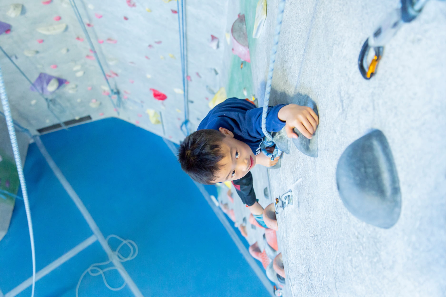 A young boy looks up the wall as he climbs on top rope at Central Rock Gym. He's reaching up high with his hand to grab the next hold.