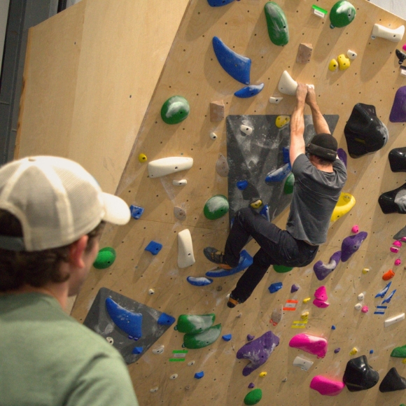 A man watches a climber who is nearing the top of a boulder.