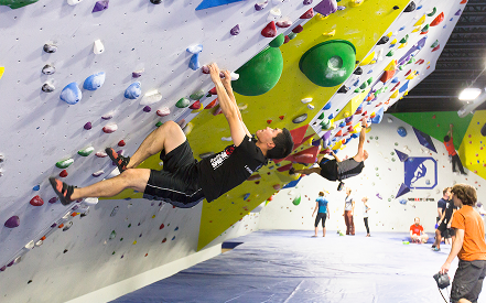 People practice indoor rock climbing on a colorful bouldering wall. Climber in foreground grips holds on a steep section, while others watch.