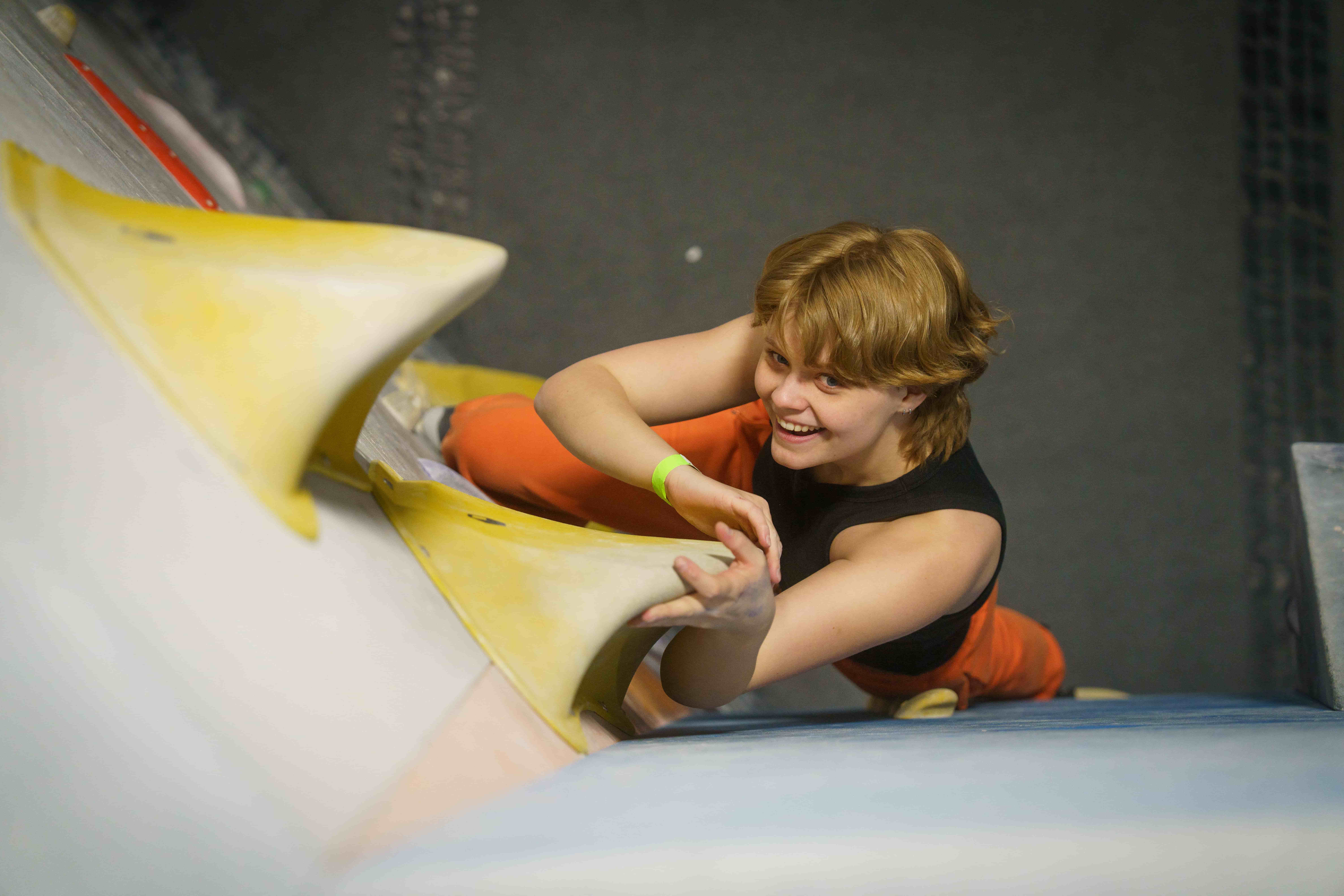 A teen girl with short hair looks up at the camera while bouldering.