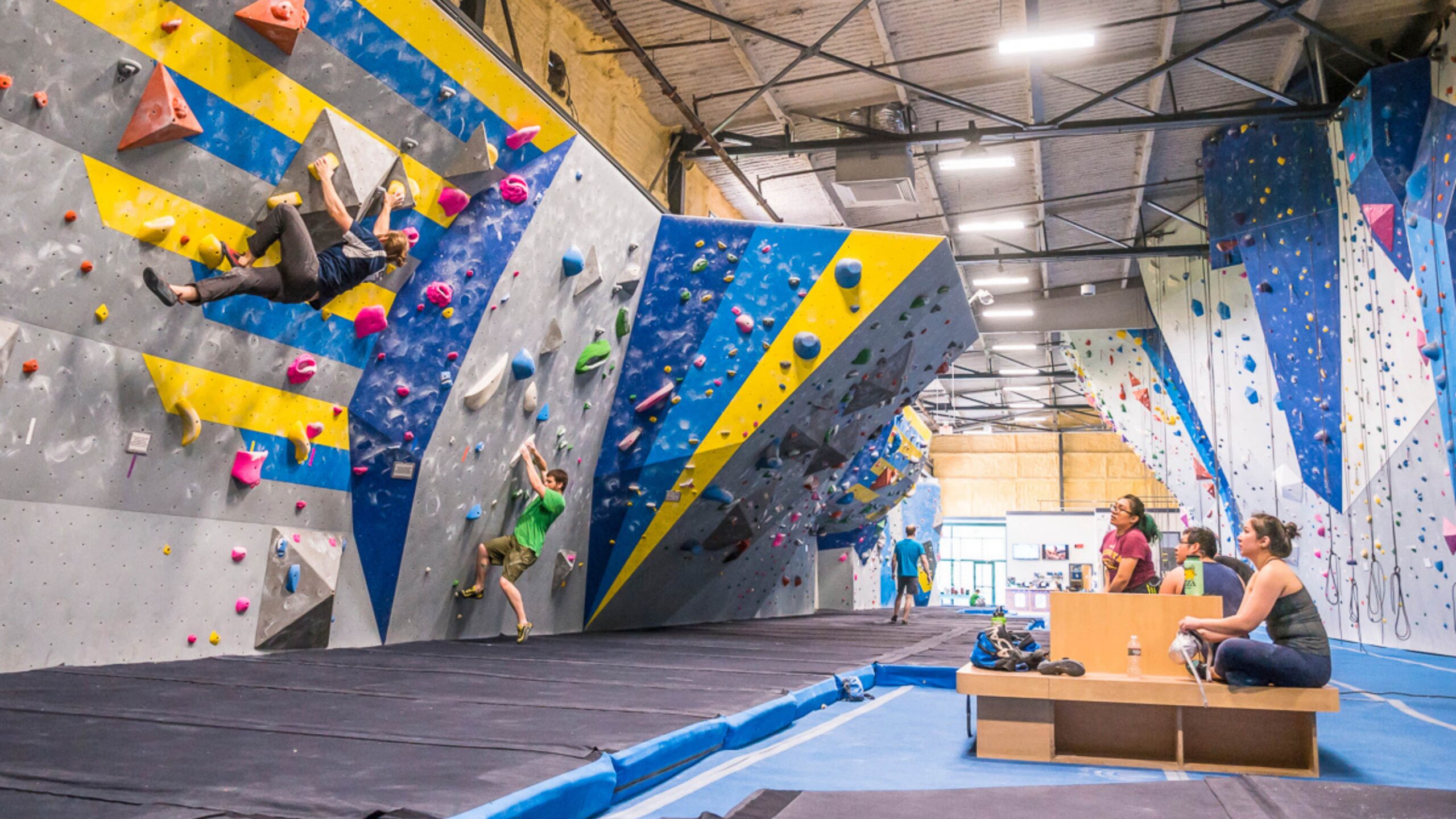 A group of climbers sit on the cubby bench and watch a man boulder and throw a backflag.