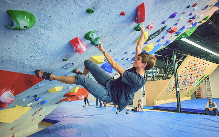 A person climbs an indoor bouldering wall with colorful holds. Others watch from padded blue mats. The scene conveys focus and athleticism.
