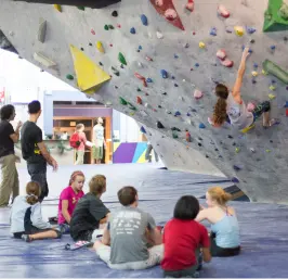 A group of kids sit on the floor watching a peer climb the bouldering wall as an instructor stands nearby at Central Rock Gym Watertown.