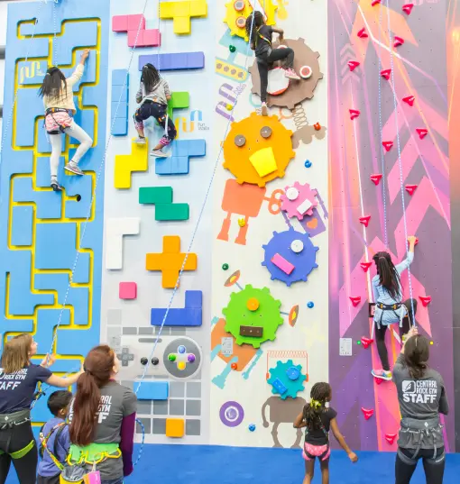 A group of kids and parents gather at the colorful children's climbing wall at Central Rock Gym Watertown during a group event, a child scaling the pink wall as a staff member looks on.