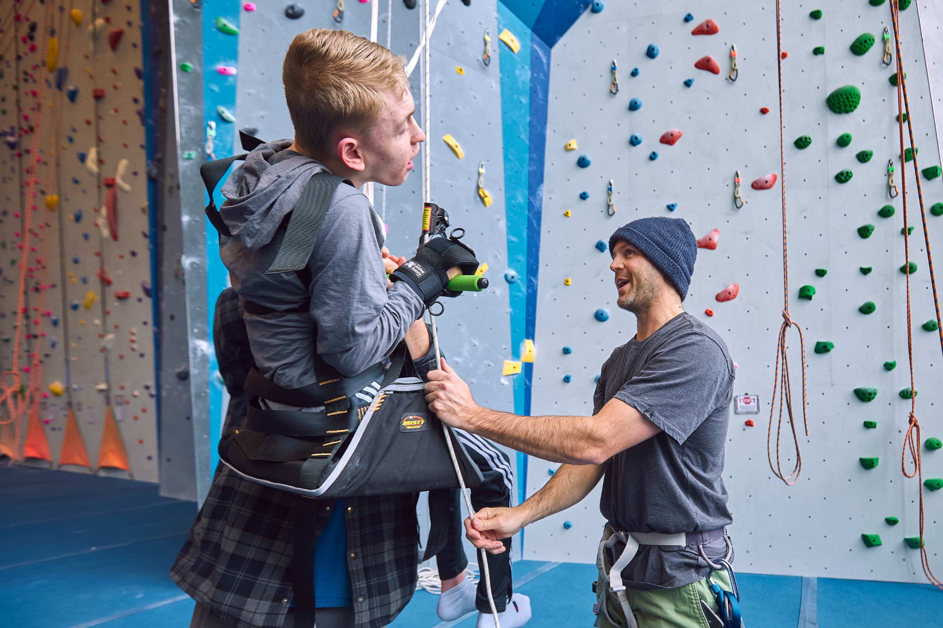 A volunteer wearing a beanie and climbing harness helps an adaptive climber get started as he sits in a seat harness and uses a pullup bar to climb the wall.