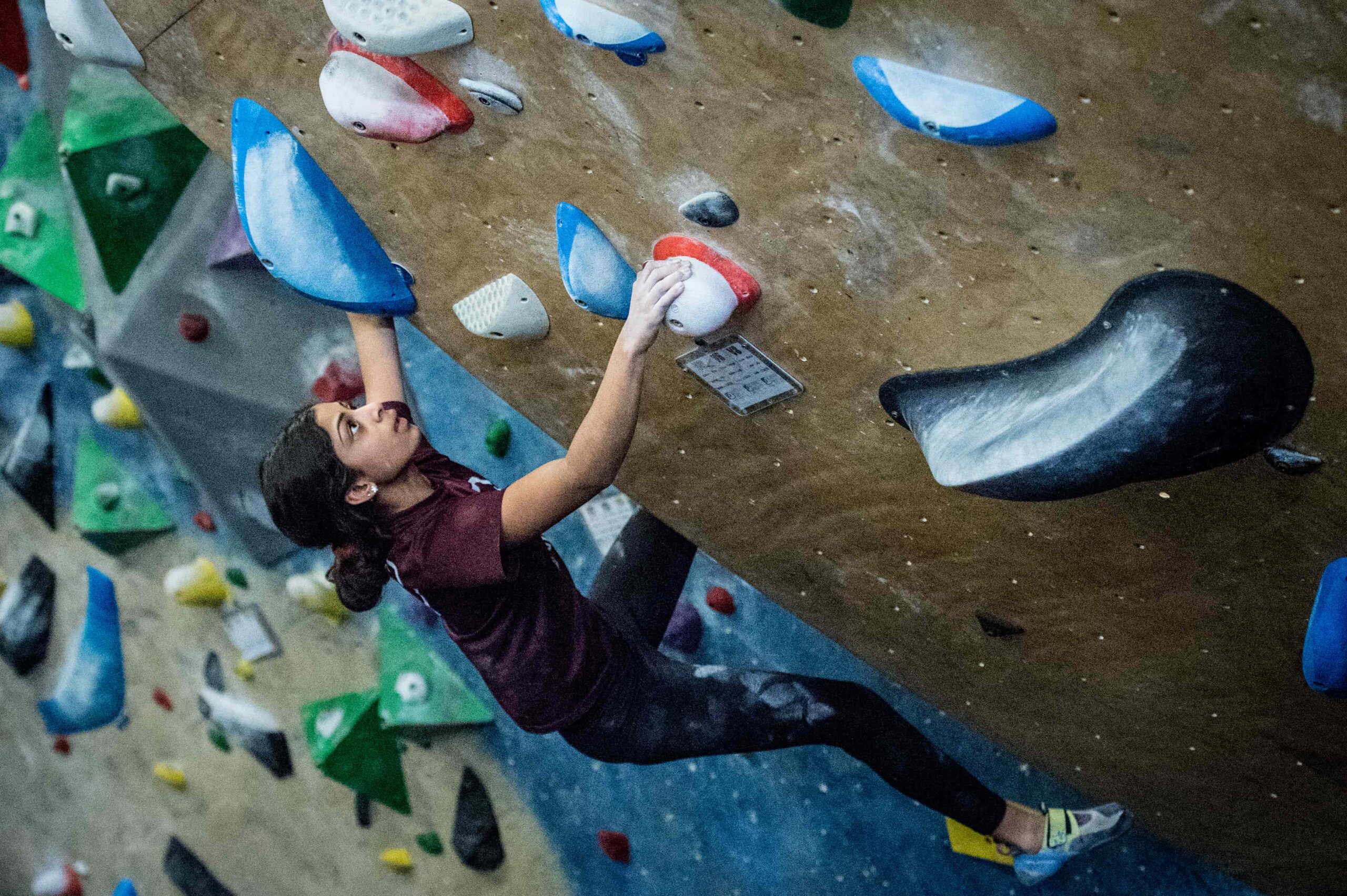 A teen girl boulders on a steep part of the wall and is looking at the next hold as she prepares to reach for it.