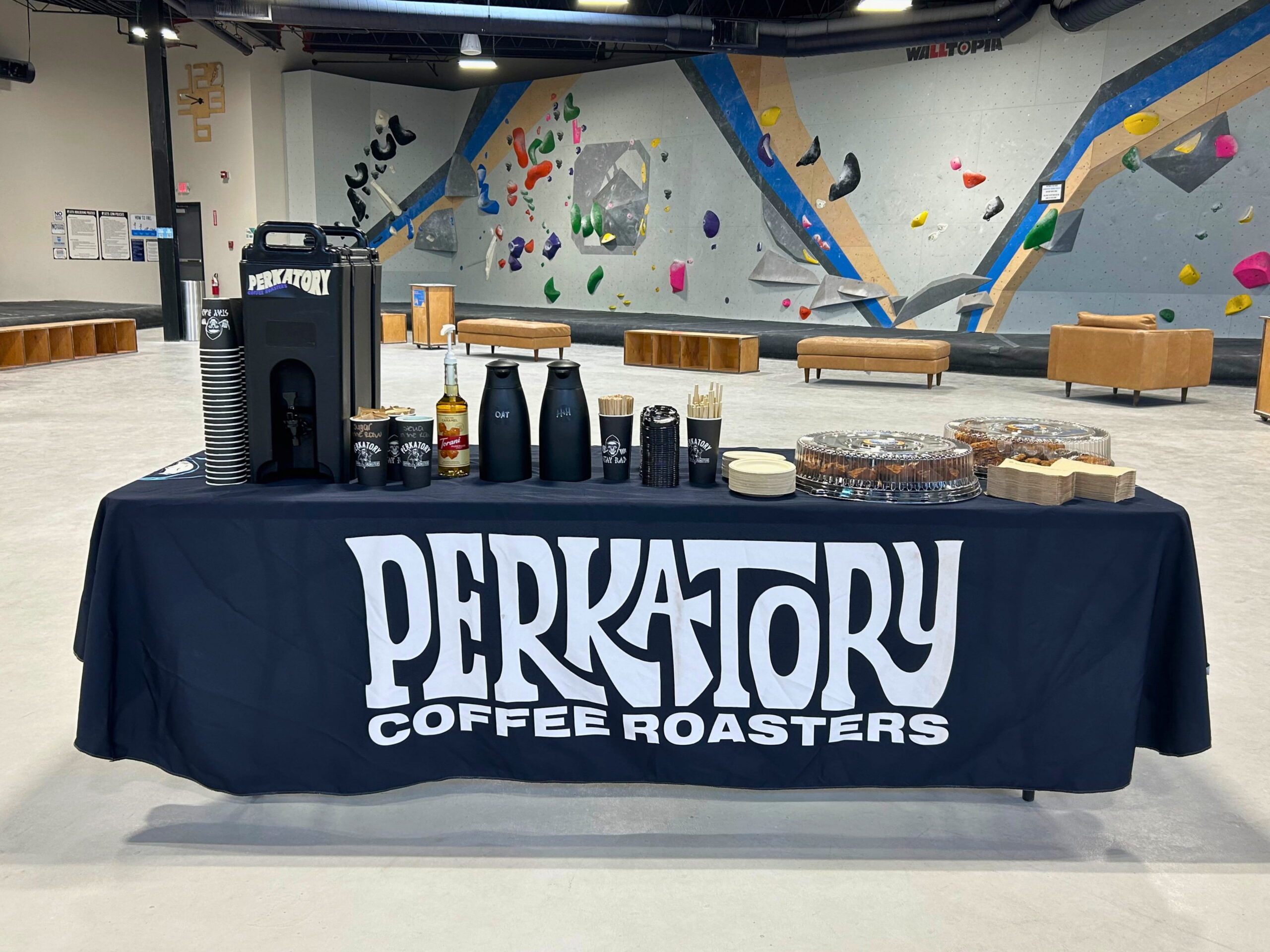 A table set up with takeaway coffee and pastries. The tablecloth reads "Perkatory Coffee Roasters".