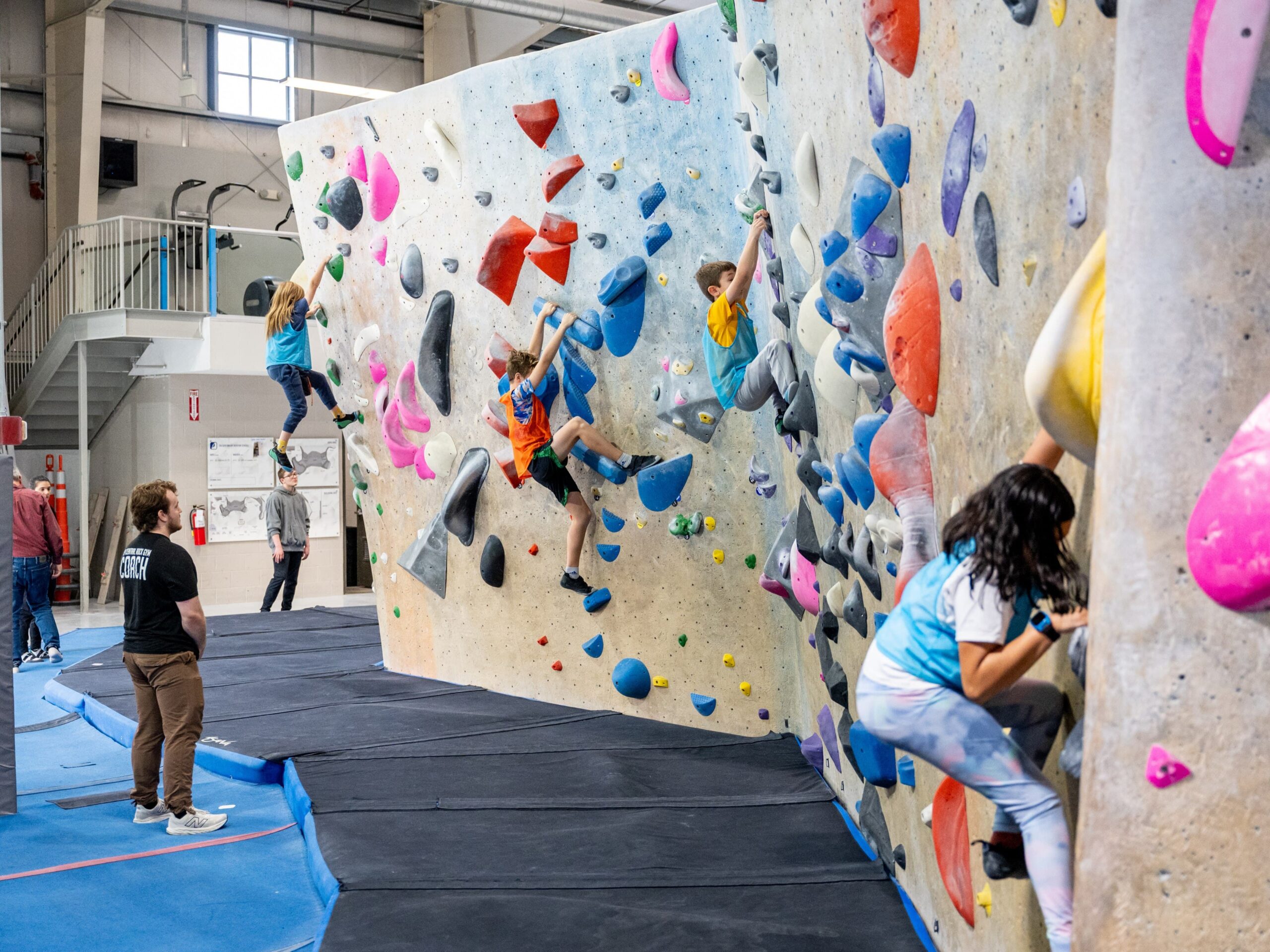 Several kids climbing on an indoor bouldering wall that features colorful holds. There is a CRG coach supervising the kids.