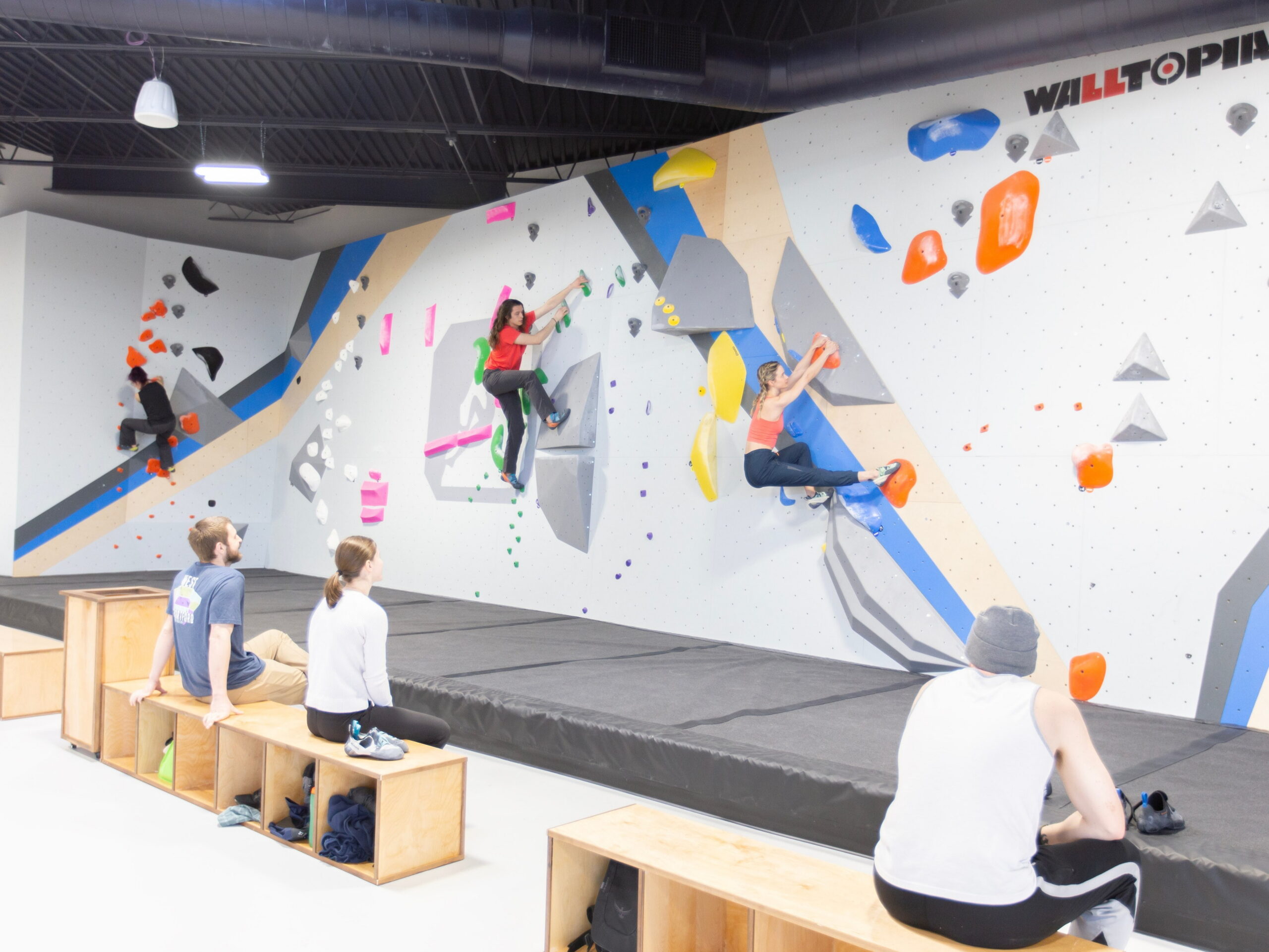 Three climbers are bouldering on an indoor rock climbing slab wall. There are three people sitting on benches watching the climbers.