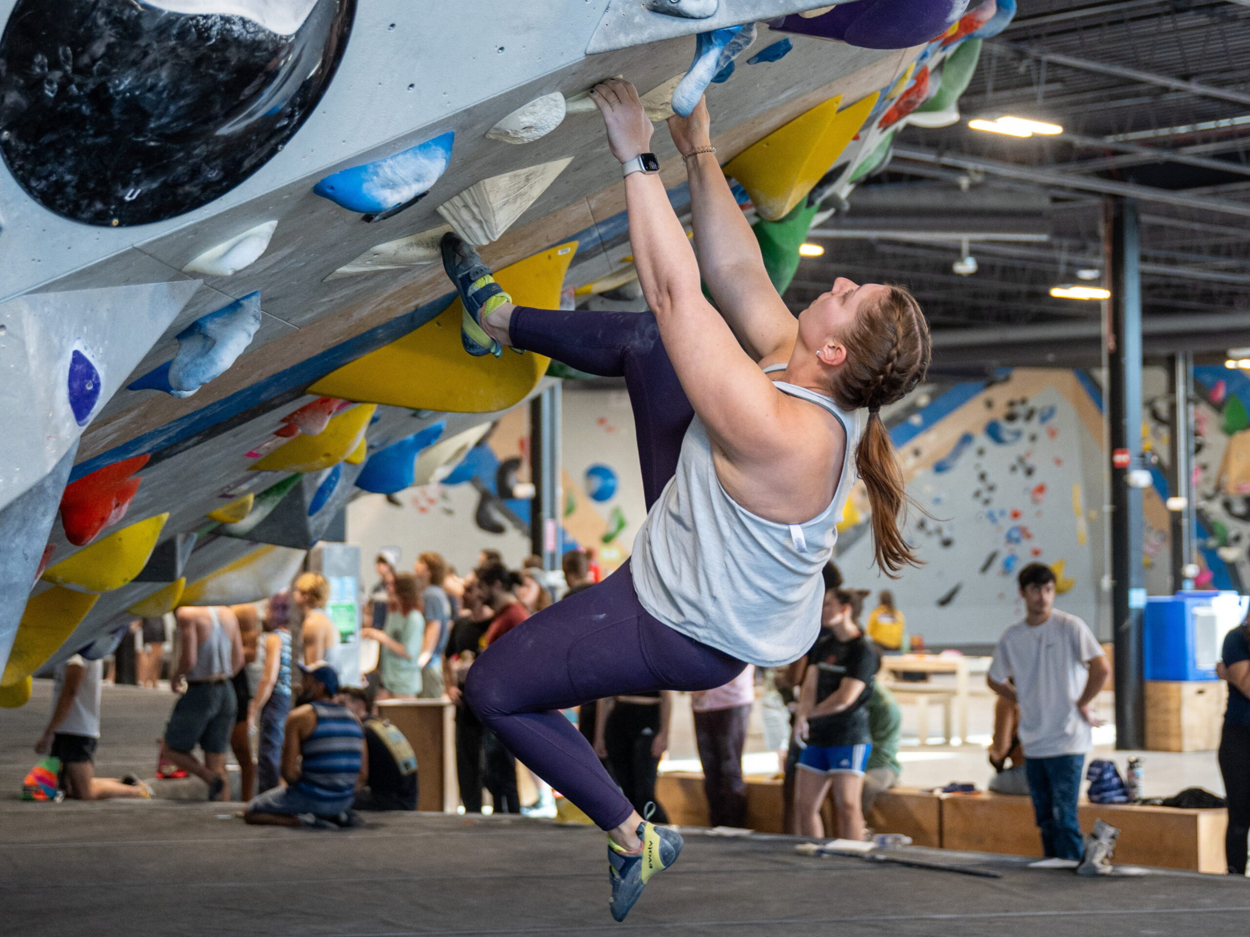 Woman bouldering on an overhanging wall at an indoor rock climbing facility. There is a crowd of people in the background.