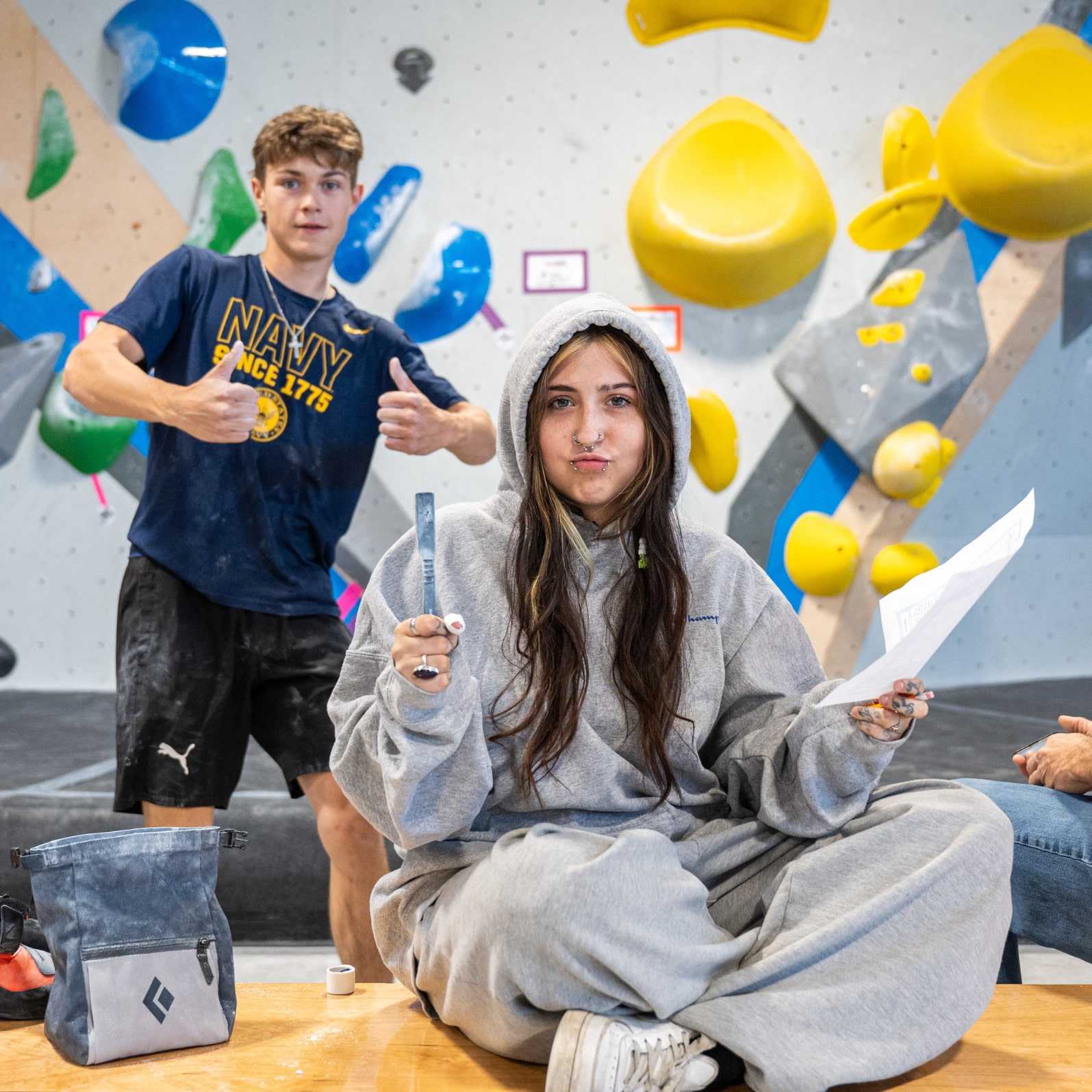Two people posing in front of an indoor bouldering wall. One person is holding a chalk brush and a piece of paper and the other person has their thumbs up.