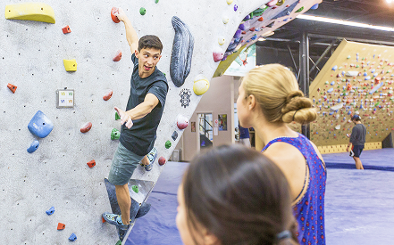A man climbs an indoor bouldering wall, reaching for a hold. Two women watch, smiling. The scene is energetic with a vibrant gym setting.