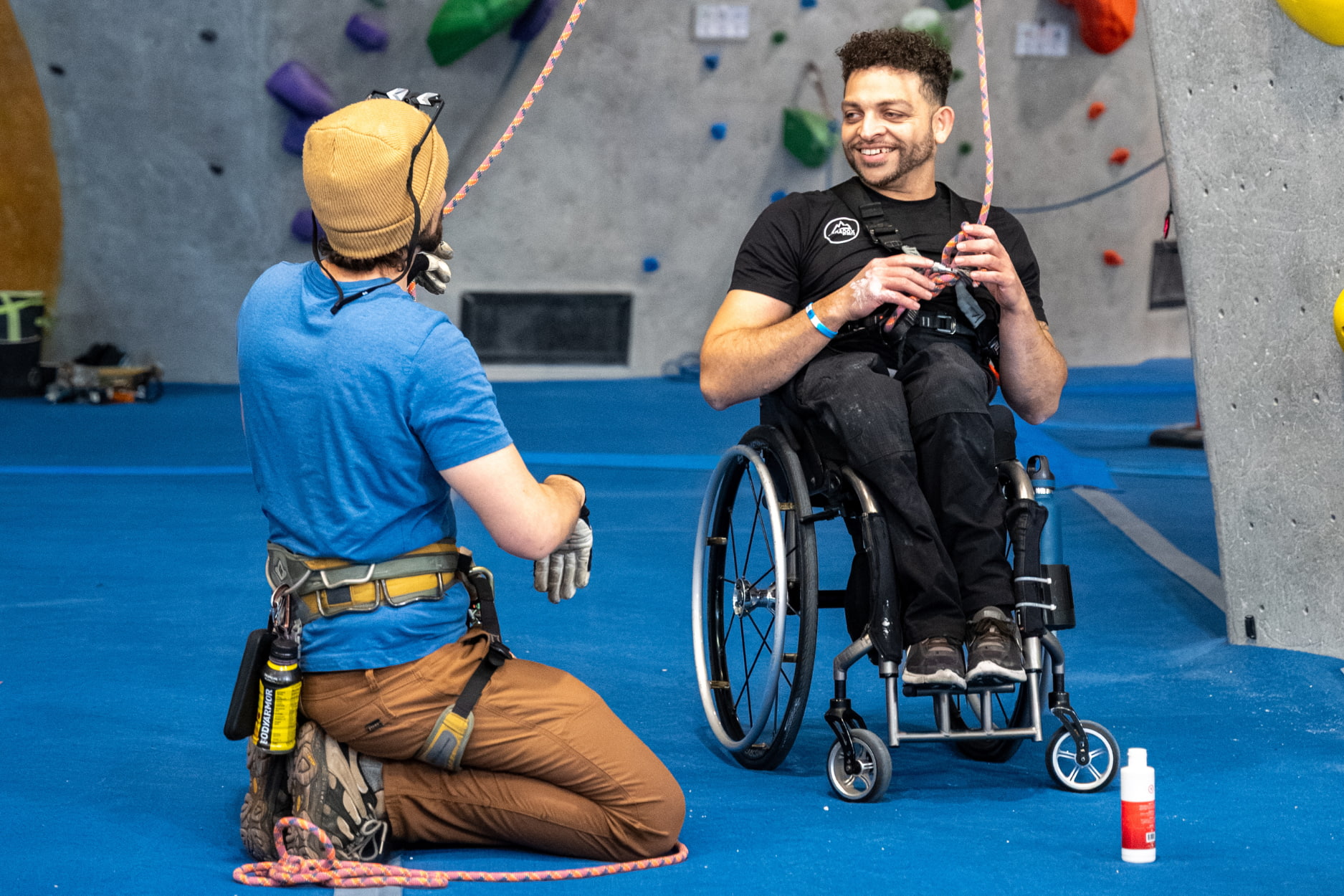 A volunteer kneels on the ground and talks to an adaptive climber in a wheel chair as he puts on his belay gloves.