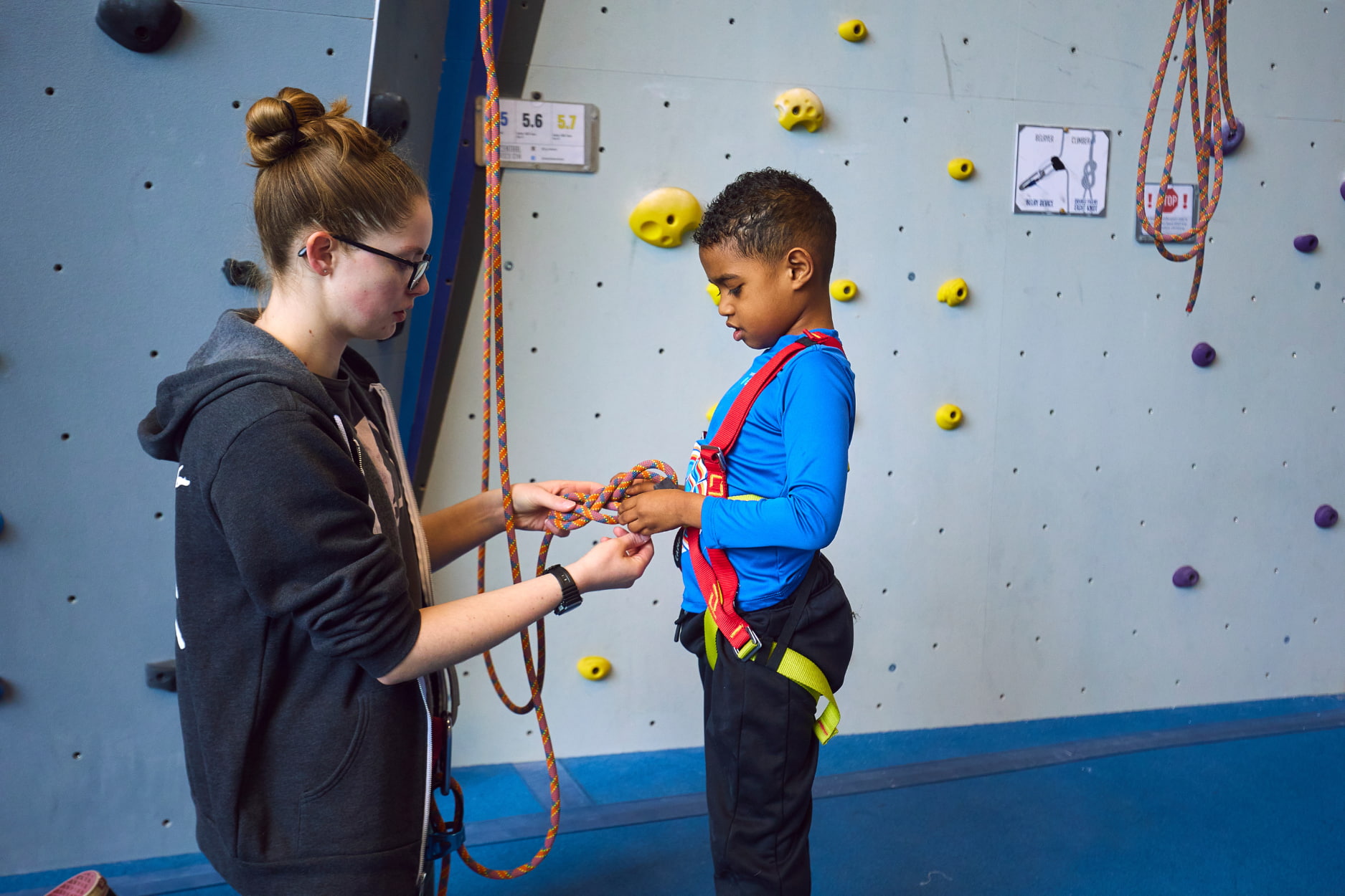A volunteer helps tie a young adaptive climber into a full body harness to begin climbing.