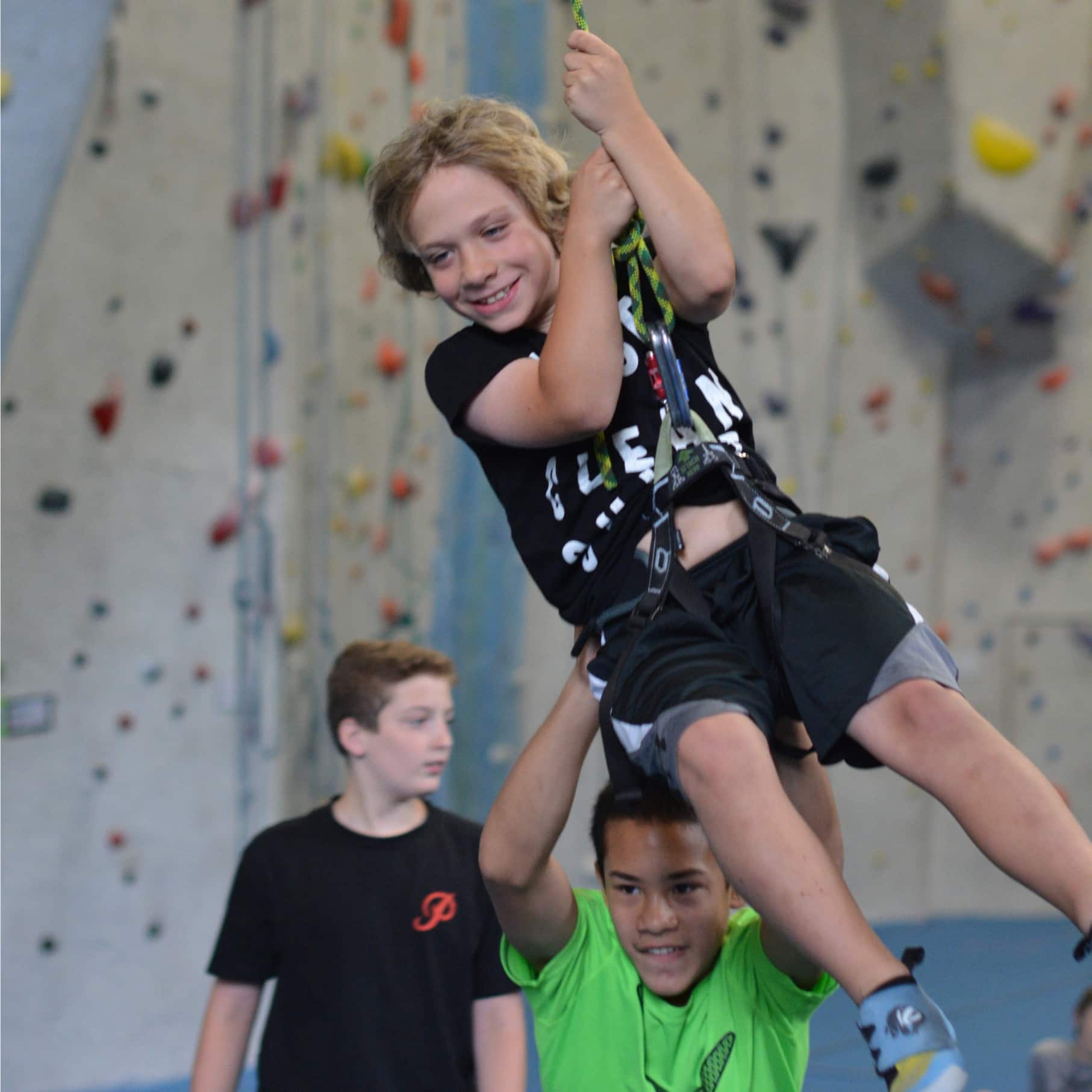 A camp kid swing on a rope at central rock gym while another kids pushes him.