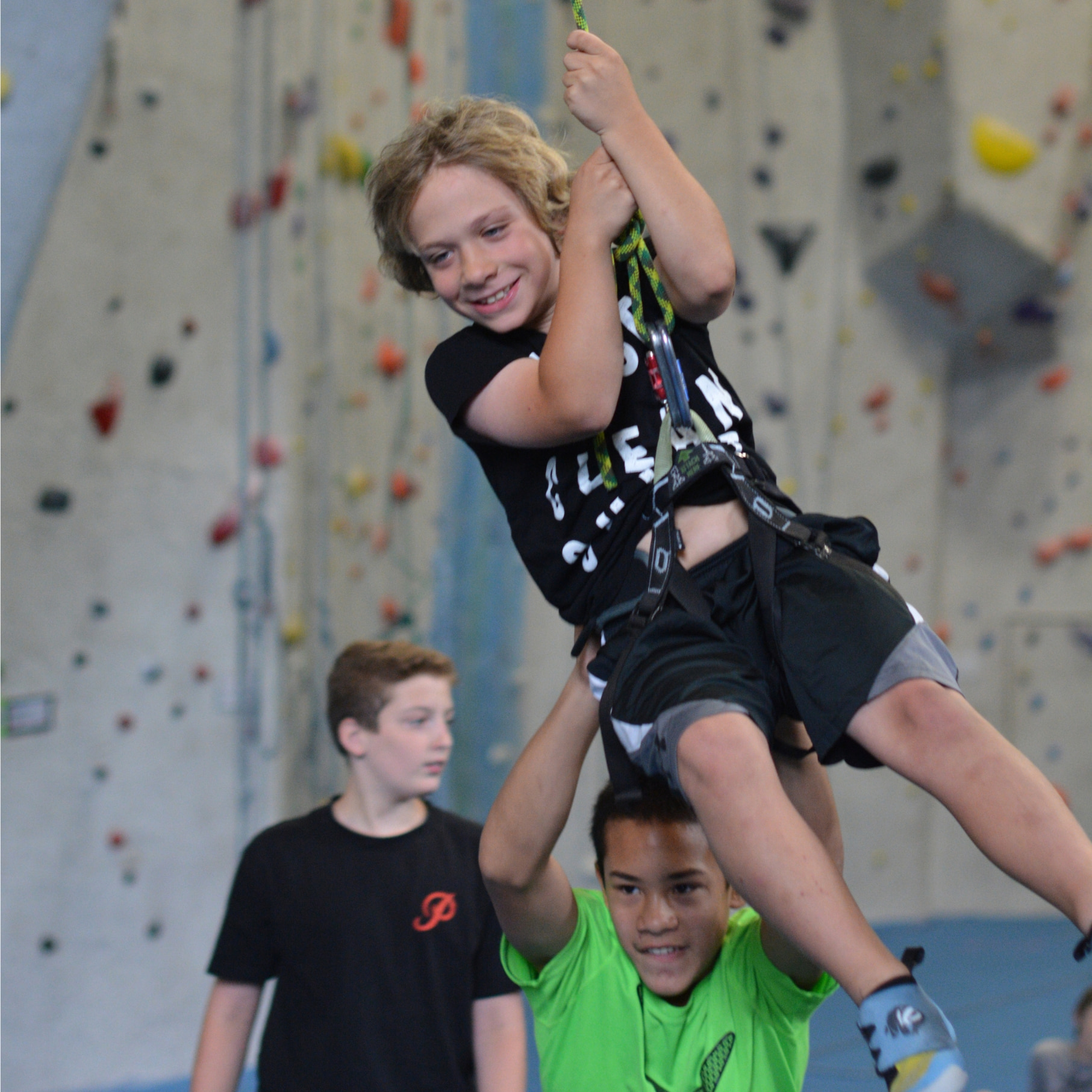 A camp kid swing on a rope at central rock gym while another kids pushes him.
