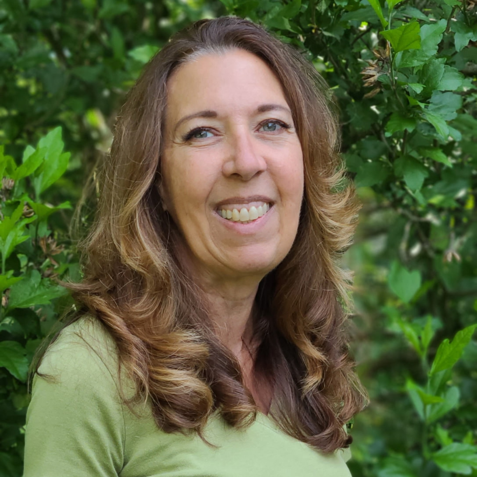 A yoga teacher with long, brown hair poses for the camera in a portrait style picture.