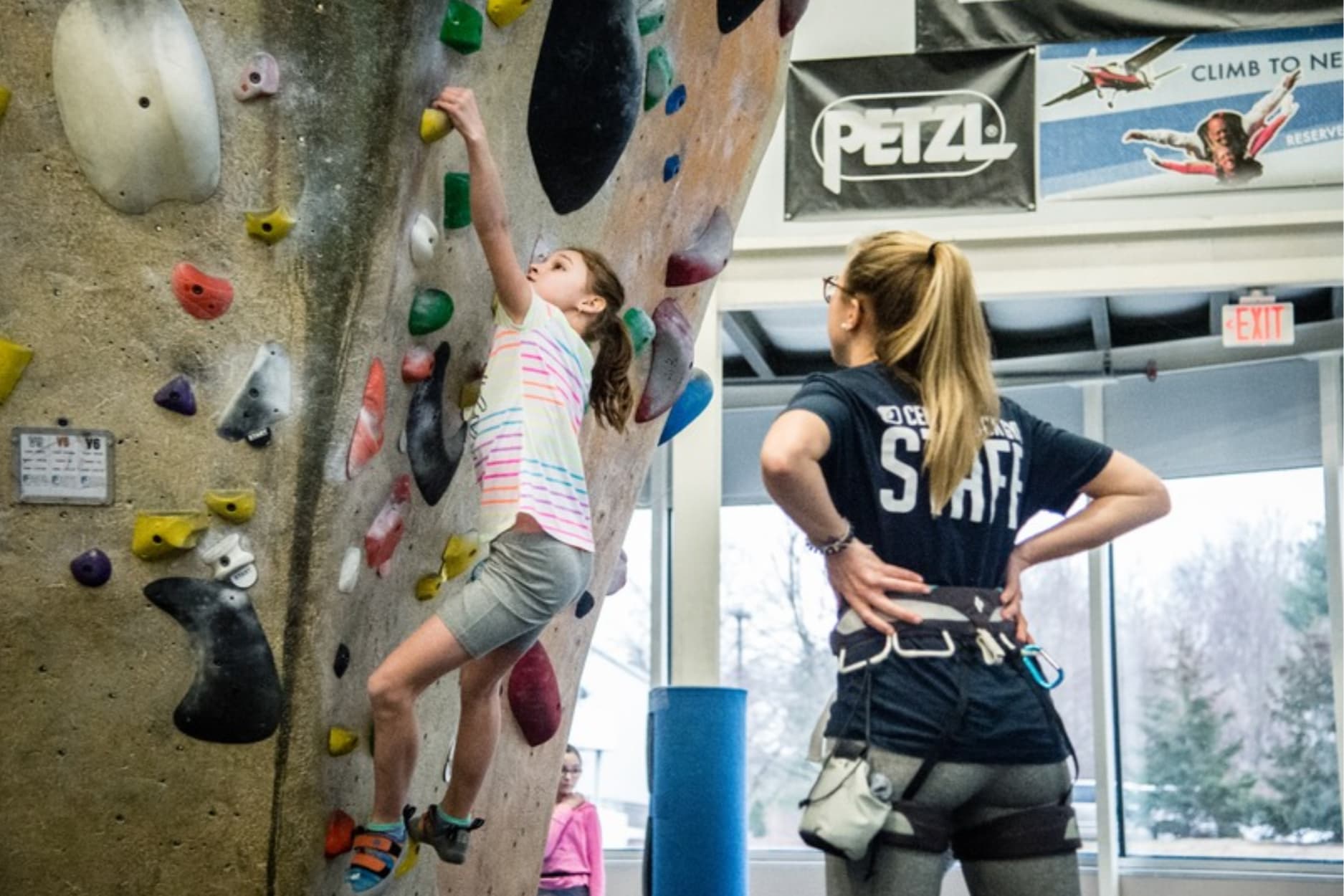 a young girl is top rope climbing while being supported by a instructor at central rock gym