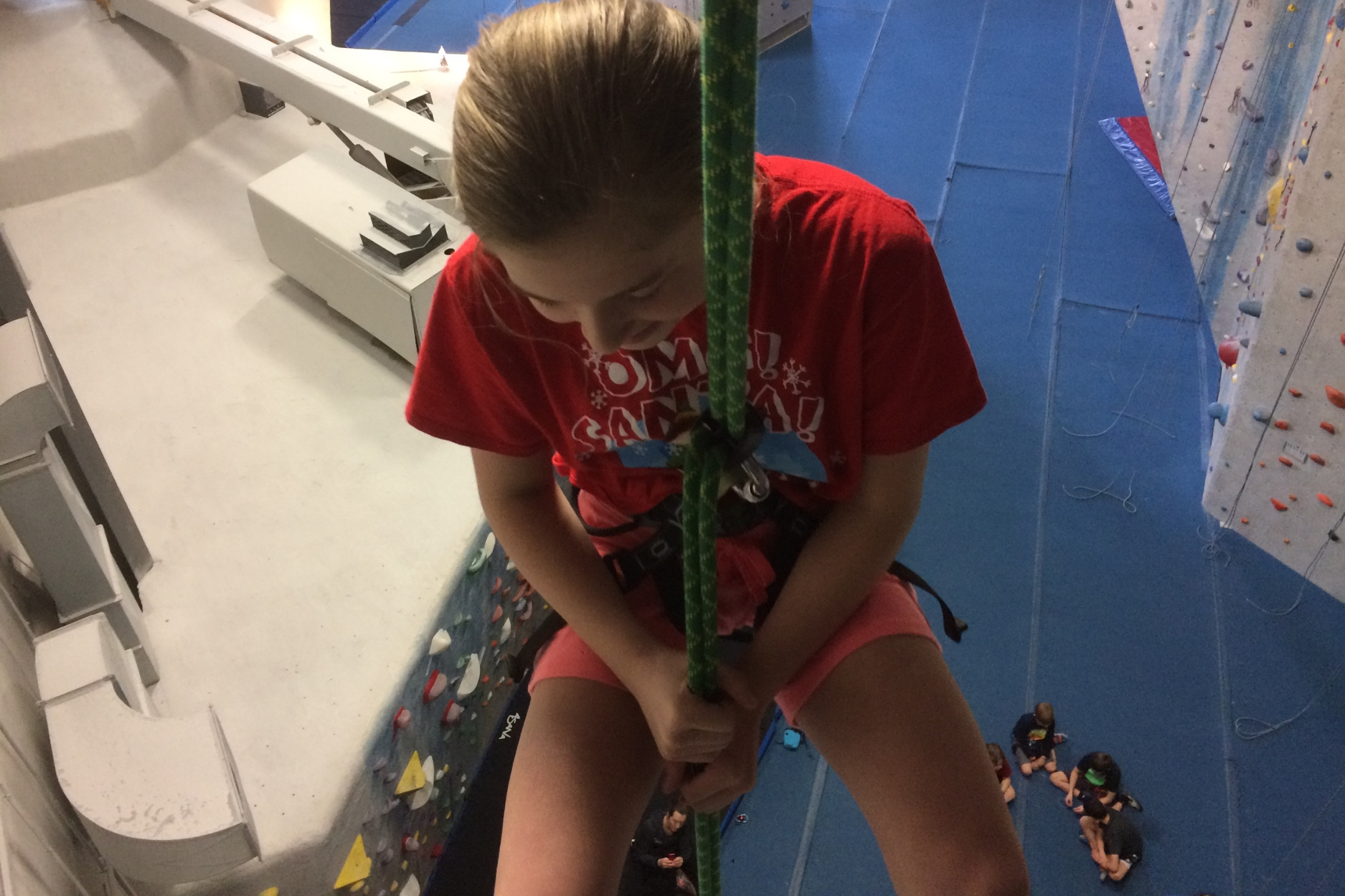 A young girl rappels from the top of a climbing wall overlooking the entire gym.
