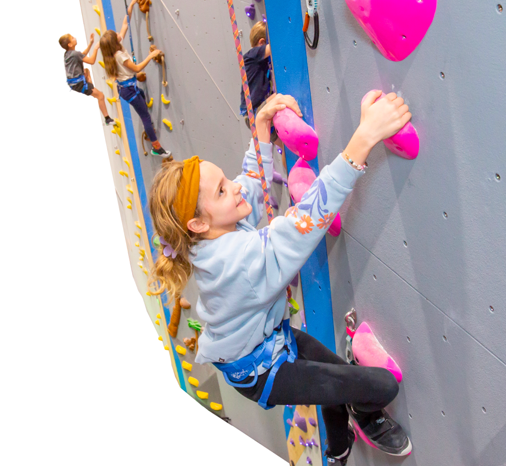 Children rock climbing indoors. The foreground features a young girl smiling as she reaches for a pink climbing hold.